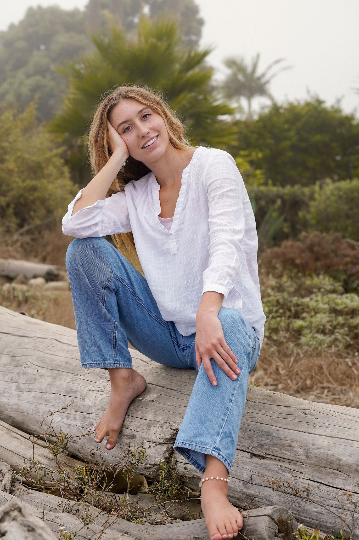 Smiling young woman with blonde hair wears a white linen shirt and jeans while seated on a driftwood log among coastal greenery.