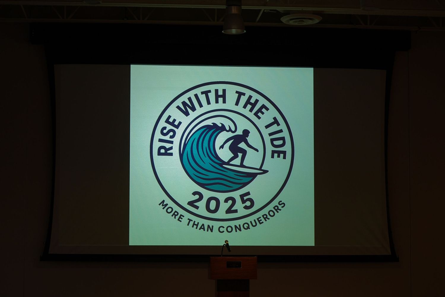 A projector screen displays the Rise with the Tide 2025 More Than Conquerors logo featuring a surfer on a wave.