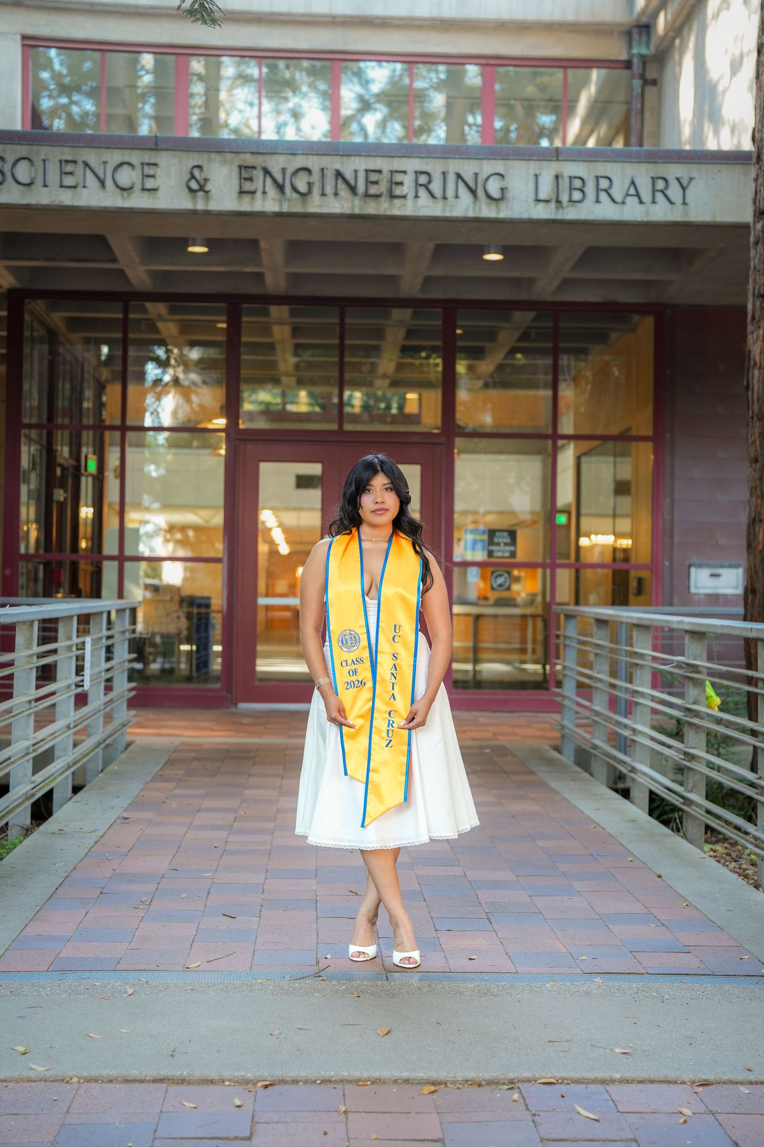Graduate in a white dress and orange sash standing on a brick walkway in front of the Science and Engineering Library.