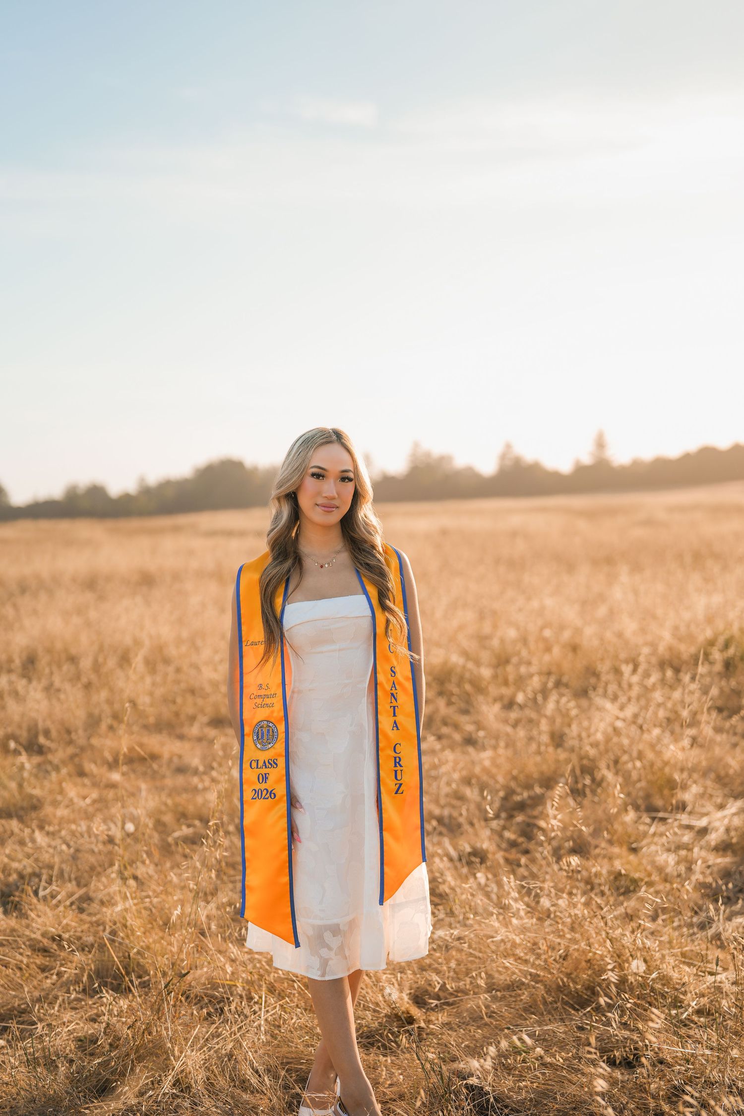 Graduate in a white dress and orange UC Santa Cruz Class of 2026 stole standing in a golden field at sunset.