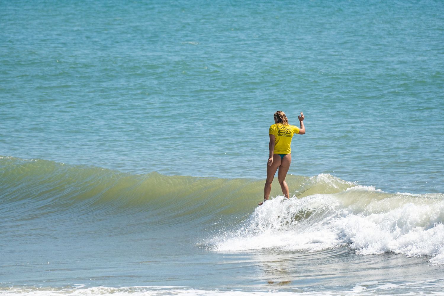 Surfer in a yellow shirt stands on a longboard with one arm raised as a wave breaks in the turquoise ocean.