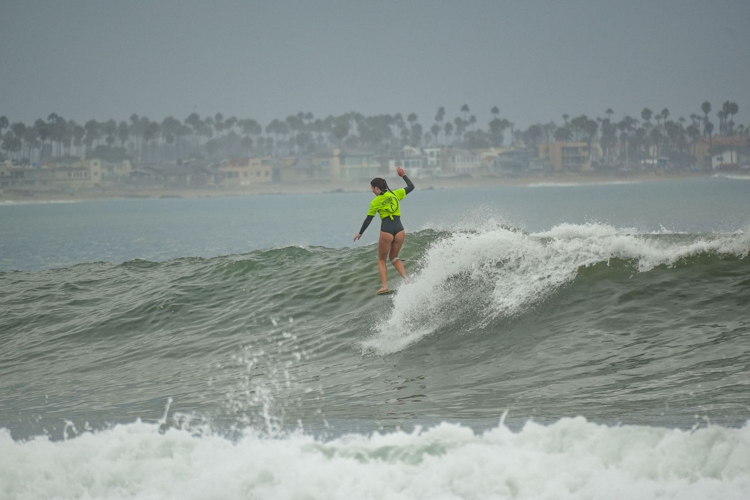 Surfer in a bright green top rides a gray-green ocean wave with a palm-lined shoreline in the background.