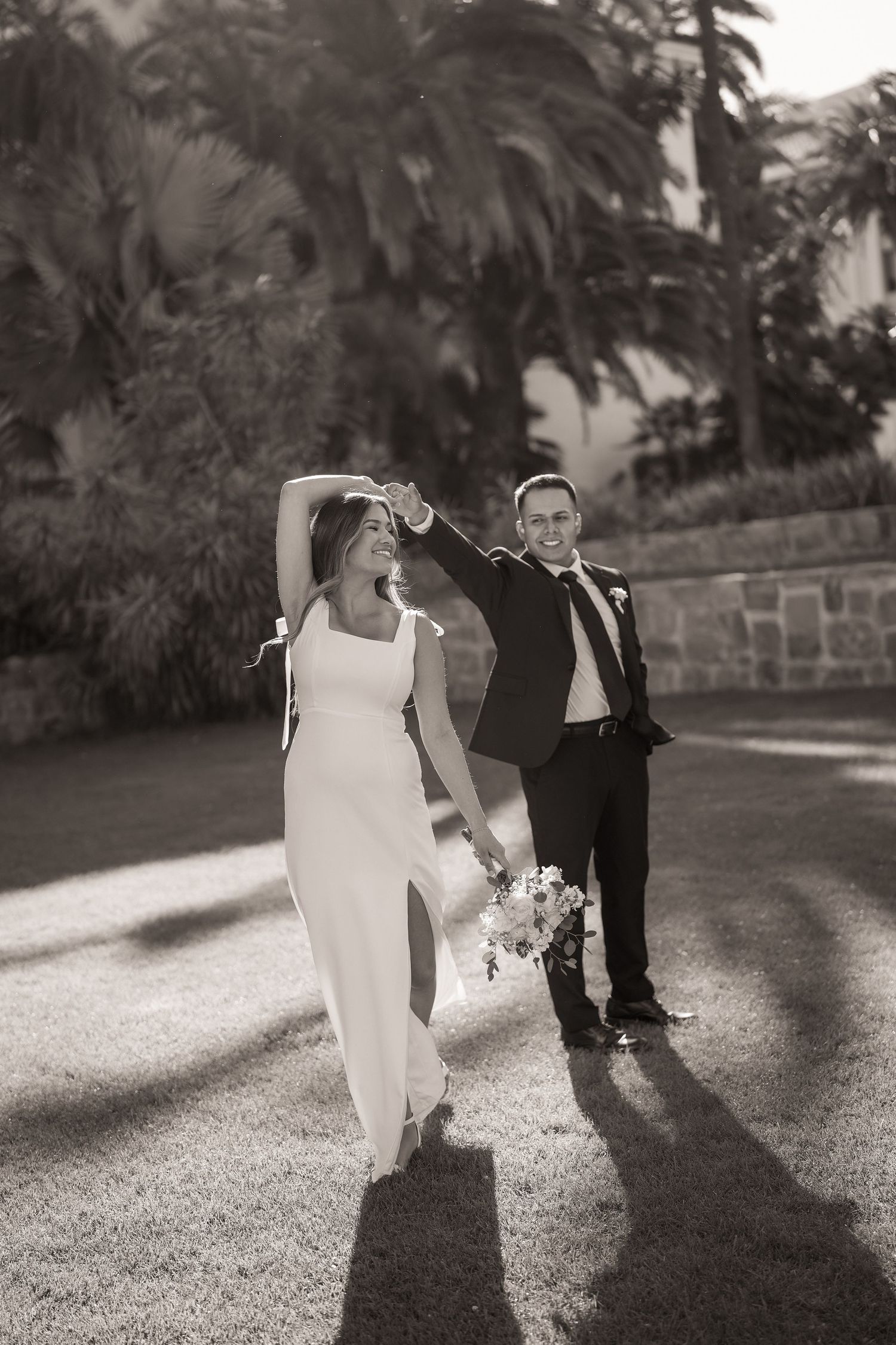 Black and white photo of a bride and groom dancing hand in hand on a sunny lawn with palm trees behind them.