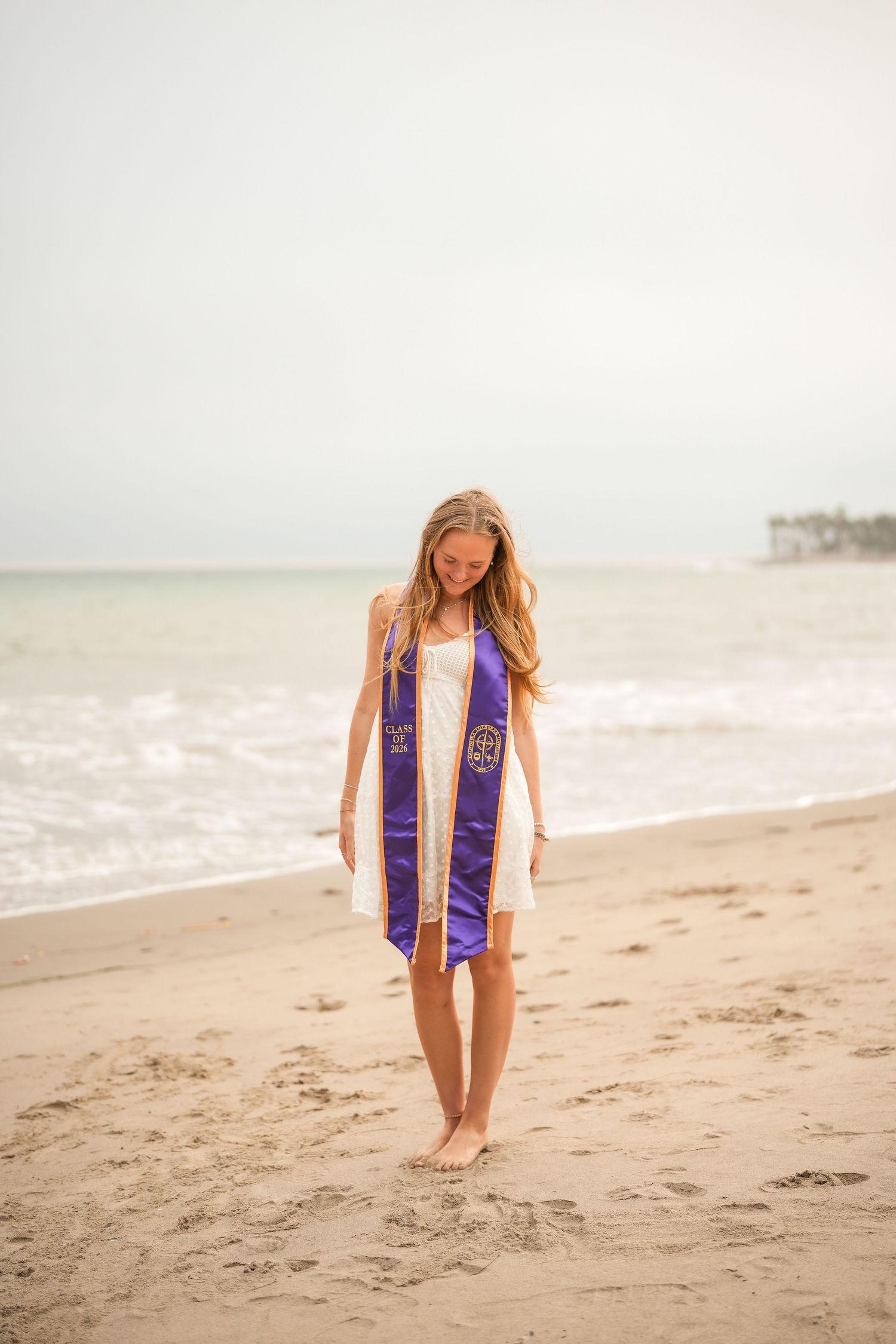 Graduate in a white dress and purple stole walks barefoot on a misty beach, smiling down at the sand.