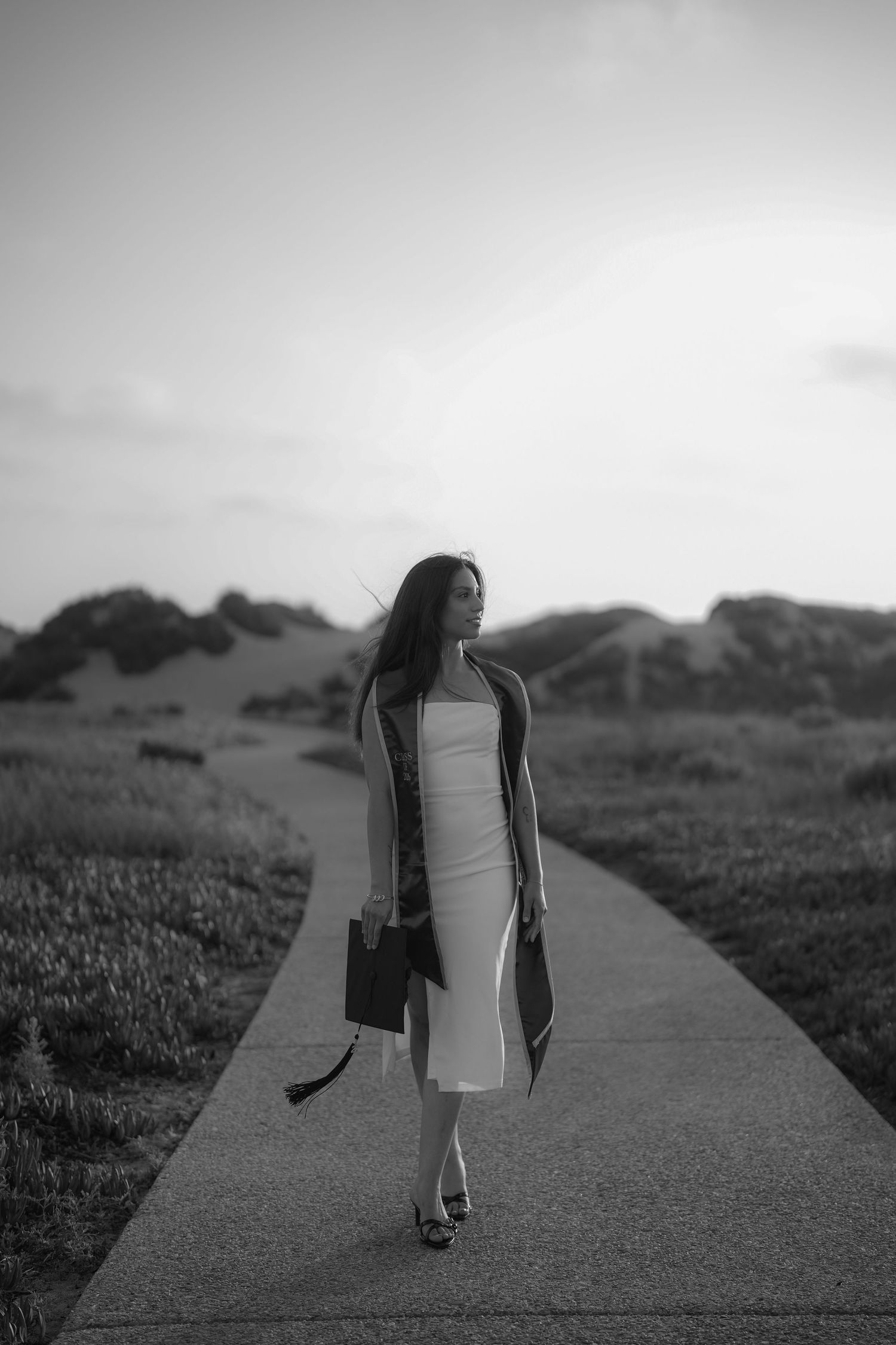 Black and white photo of a woman in a white dress and graduation sash walking along a path through grassy dunes at dusk.