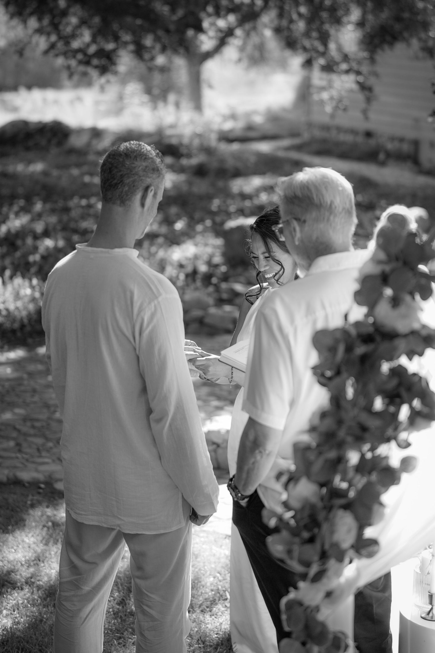 In this black and white photo, a couple exchanges rings in front of an officiant during a garden wedding ceremony with floral decor.
