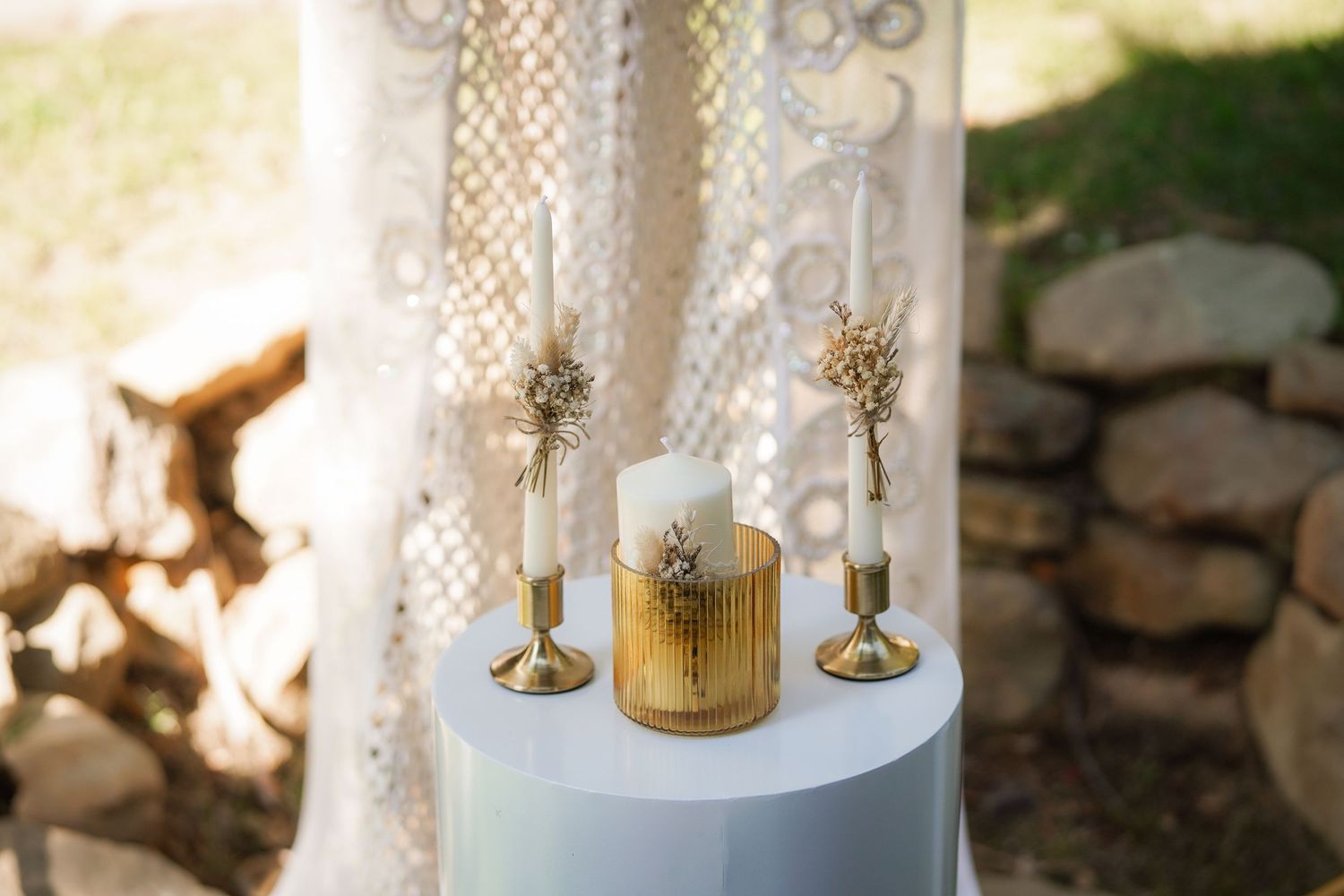 Two gold candlesticks and a lit pillar candle in a gold holder sit on a white table before lace drapes and a stone wall.