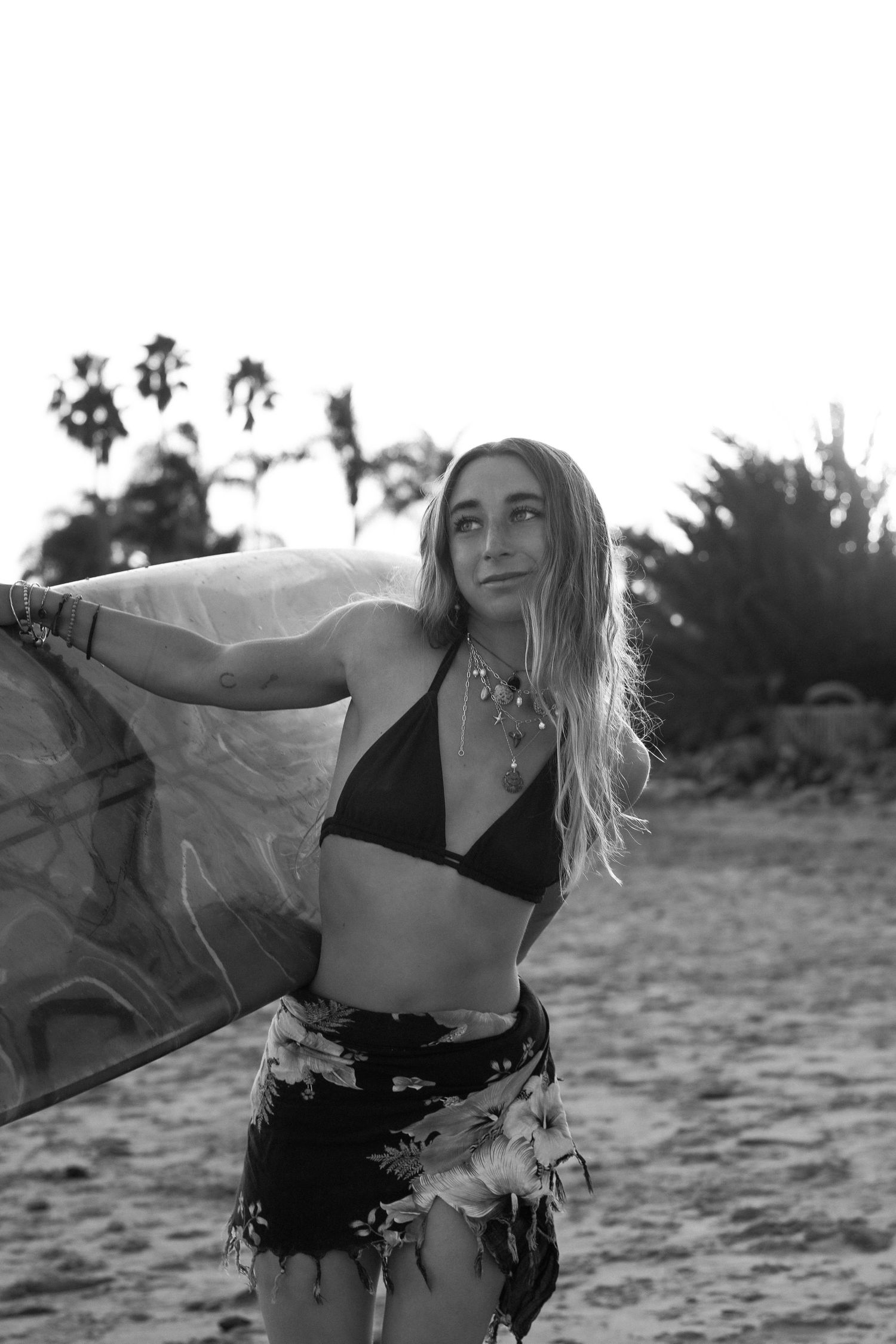 Black and white photo of a woman in a bikini top and floral skirt holding a flowing sarong on a breezy beach.