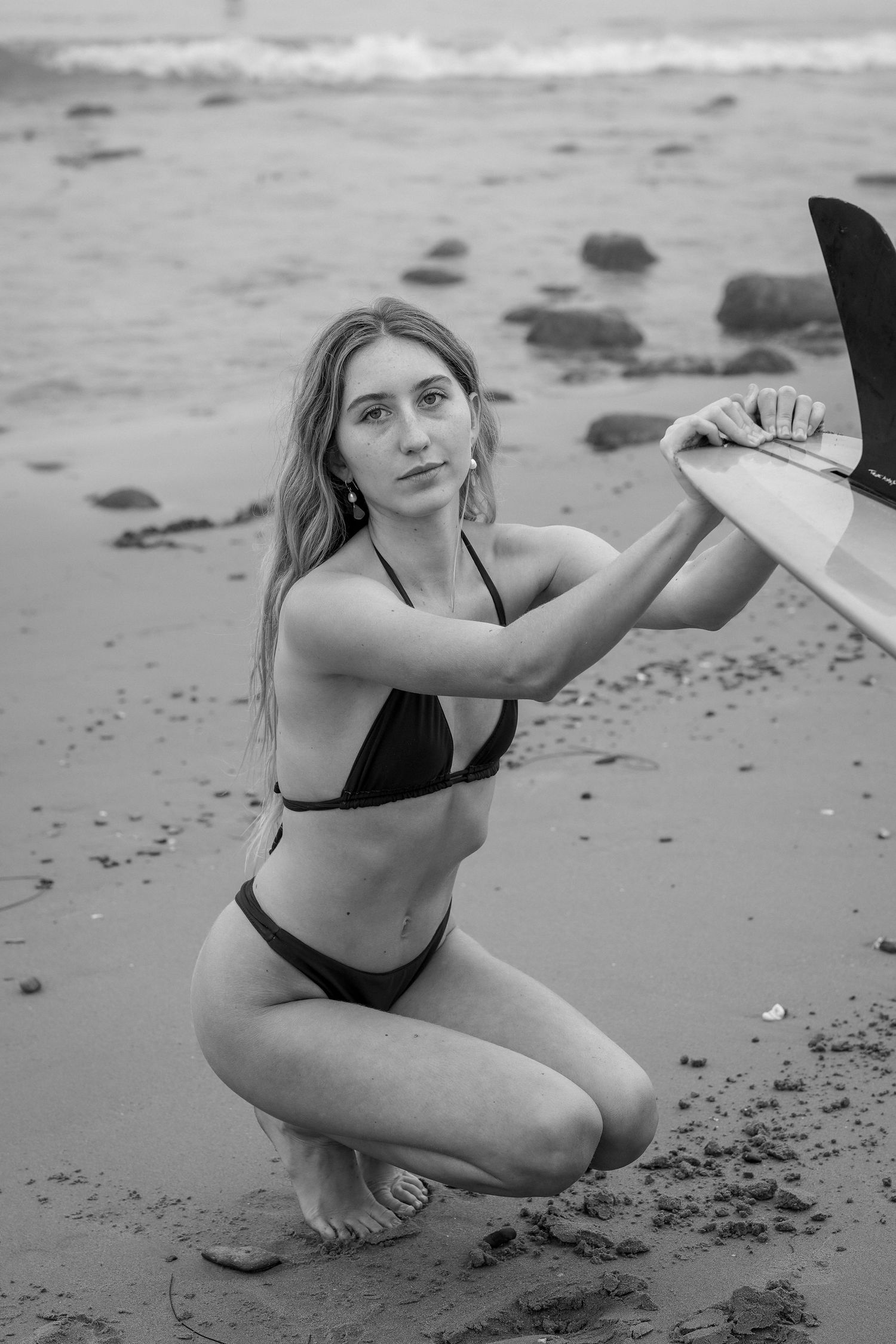 Black and white photo of a woman in a bikini crouching on a rocky beach, holding a surfboard with the ocean behind her.