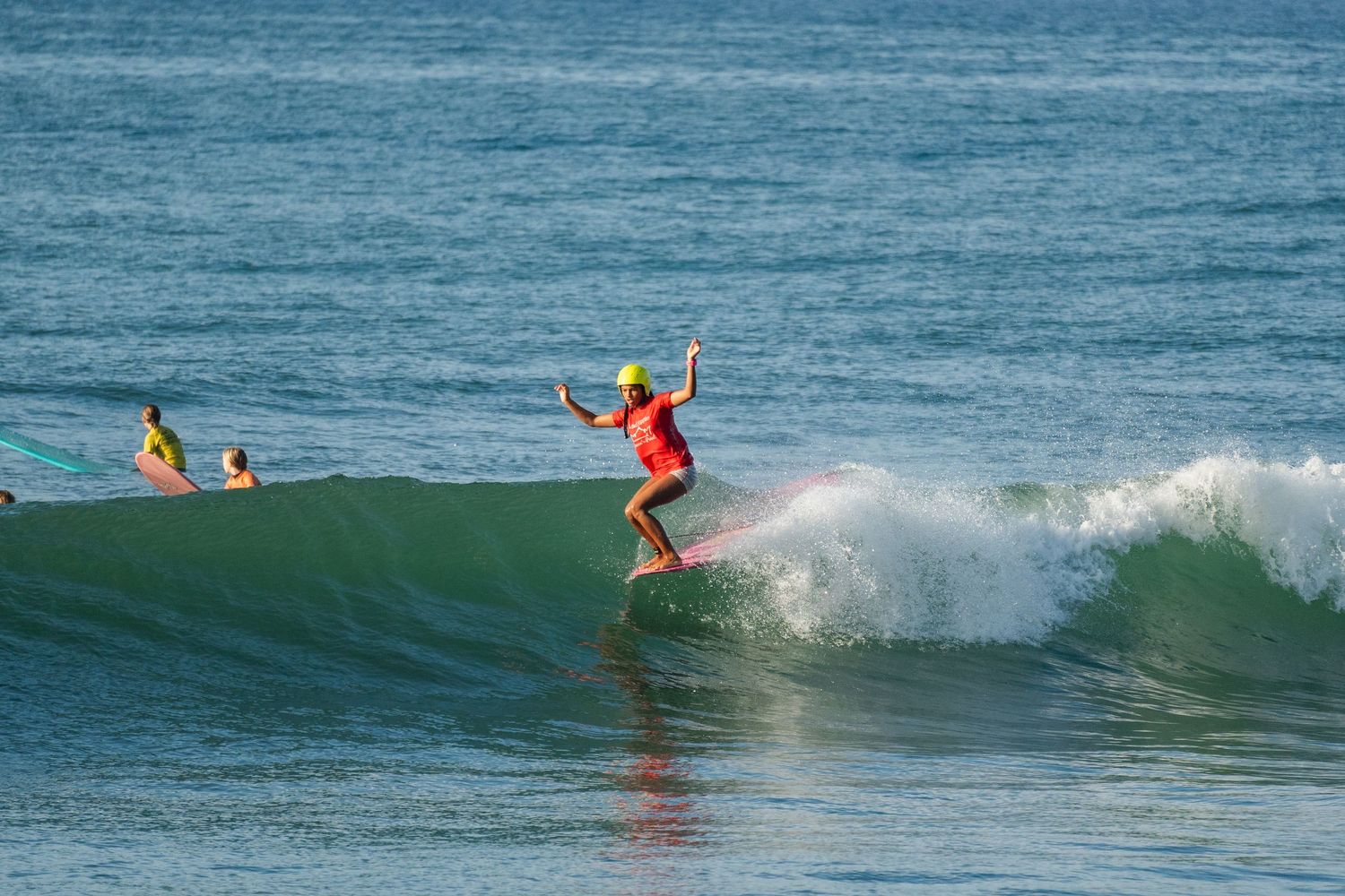 A surfer in a red top and yellow helmet balances with arms out on a pink surfboard riding a wave while two surfers watch nearby.