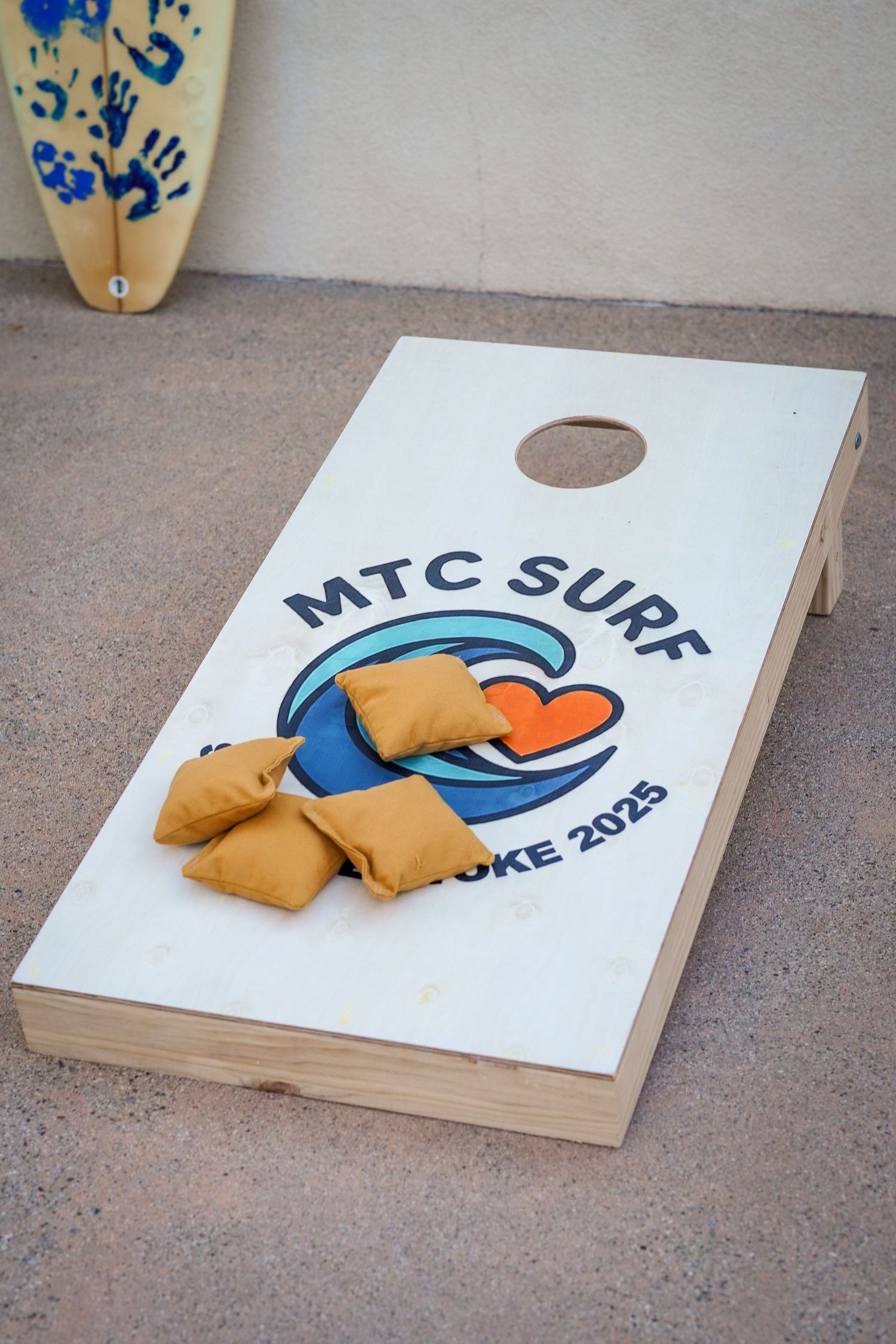MTC Surf 2025 cornhole board with orange bean bags sits on the ground beside a painted surfboard.