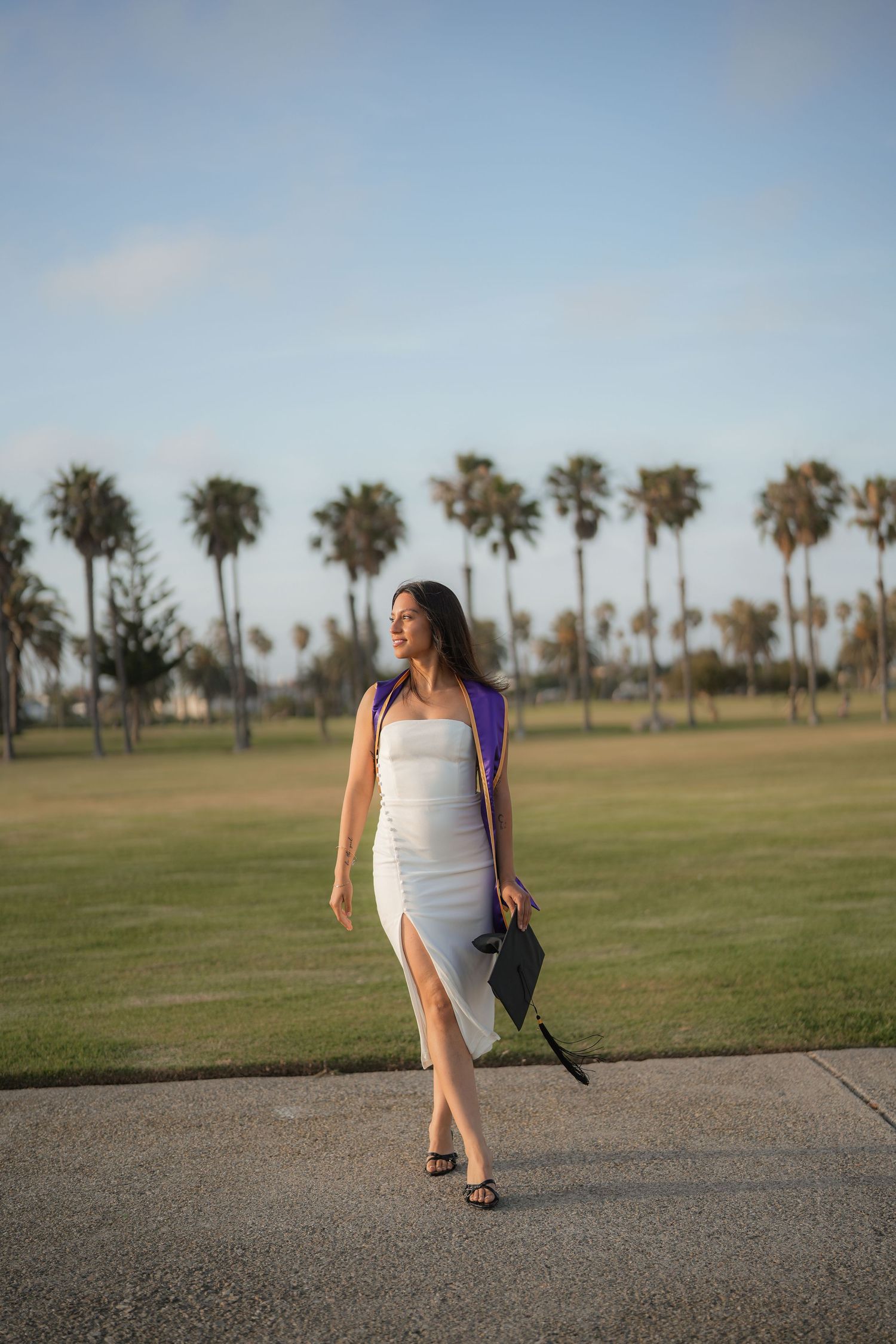 Graduate in a white slit dress with a purple stole walks on a path holding her cap, with palm trees and green lawn behind.