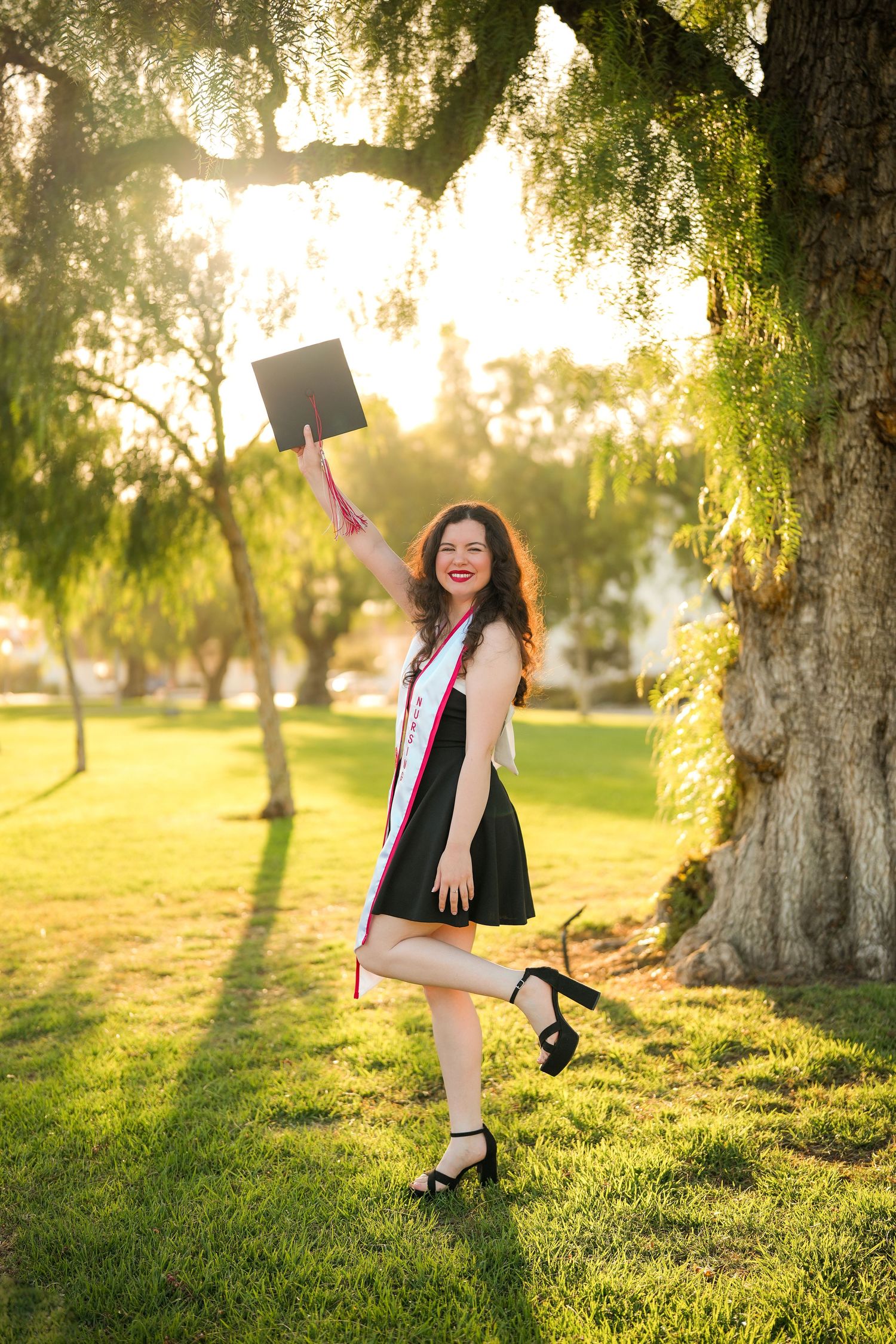 A cheerful nursing graduate in a black dress and heels tosses her cap in the air on a sunlit lawn under a large tree.