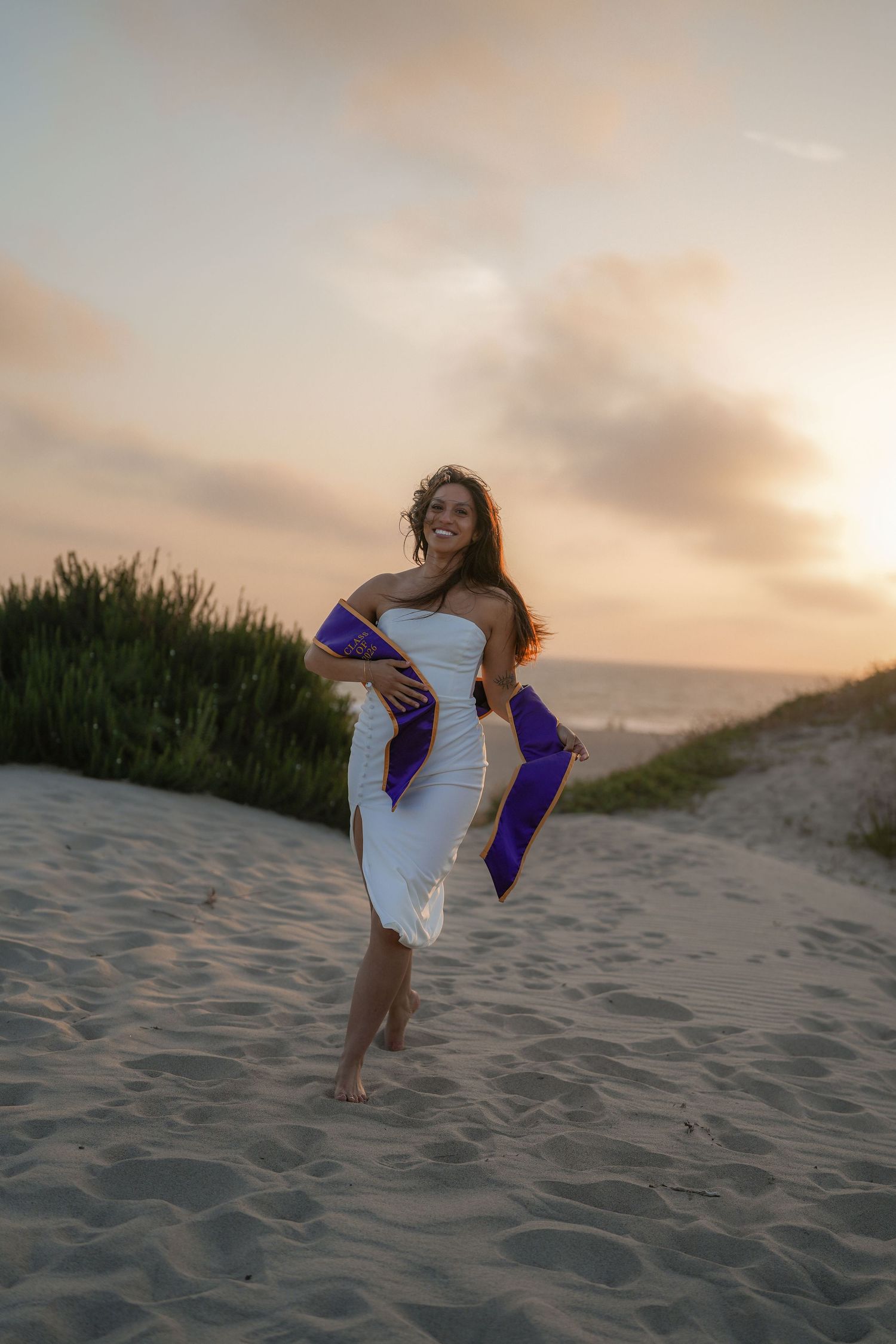 Smiling graduate in a white dress and purple sashes walking barefoot on a sandy beach at sunset.