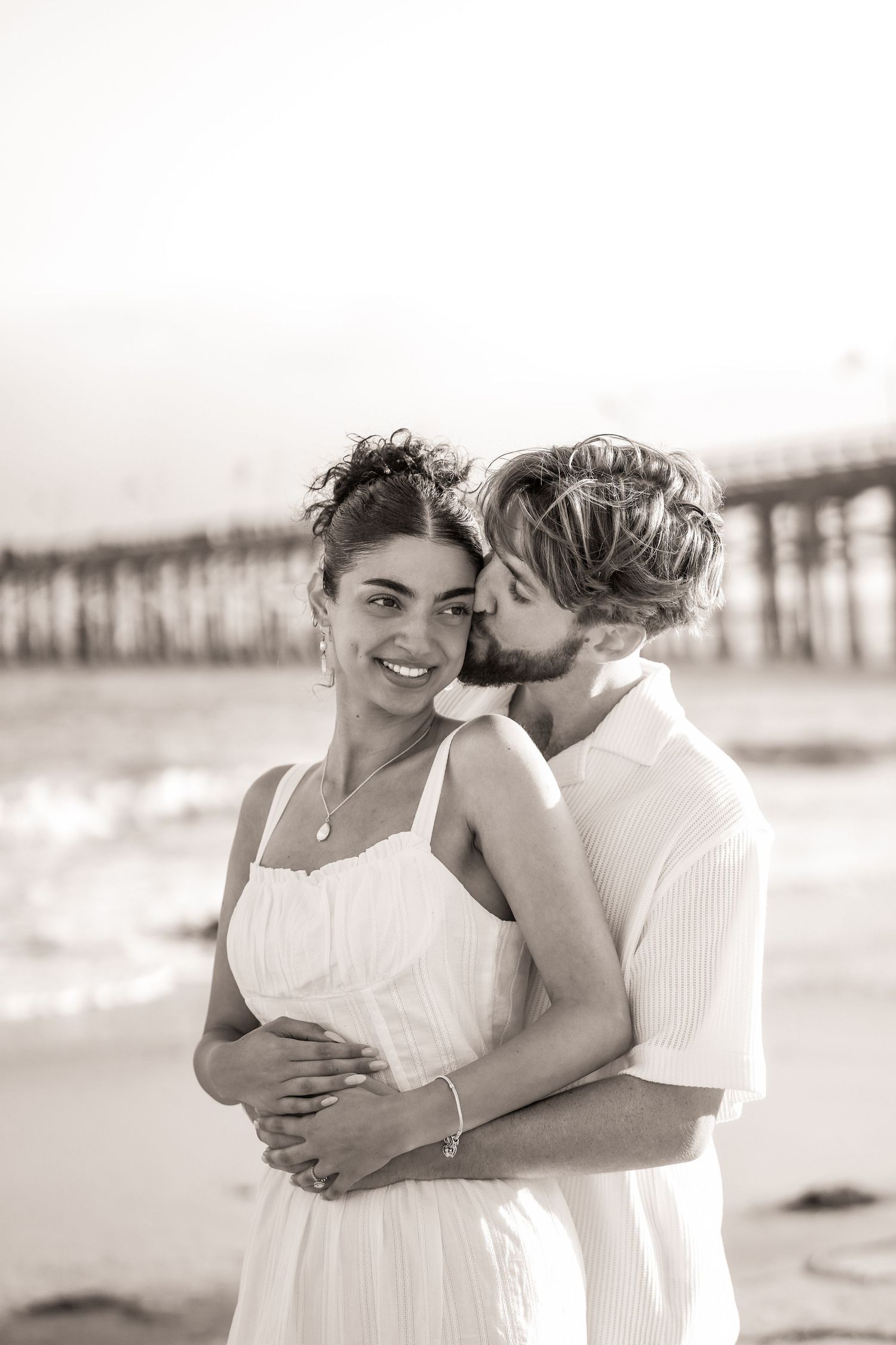 Sepia portrait of a smiling woman in a white dress as a man kisses her cheek, with a beach pier behind.