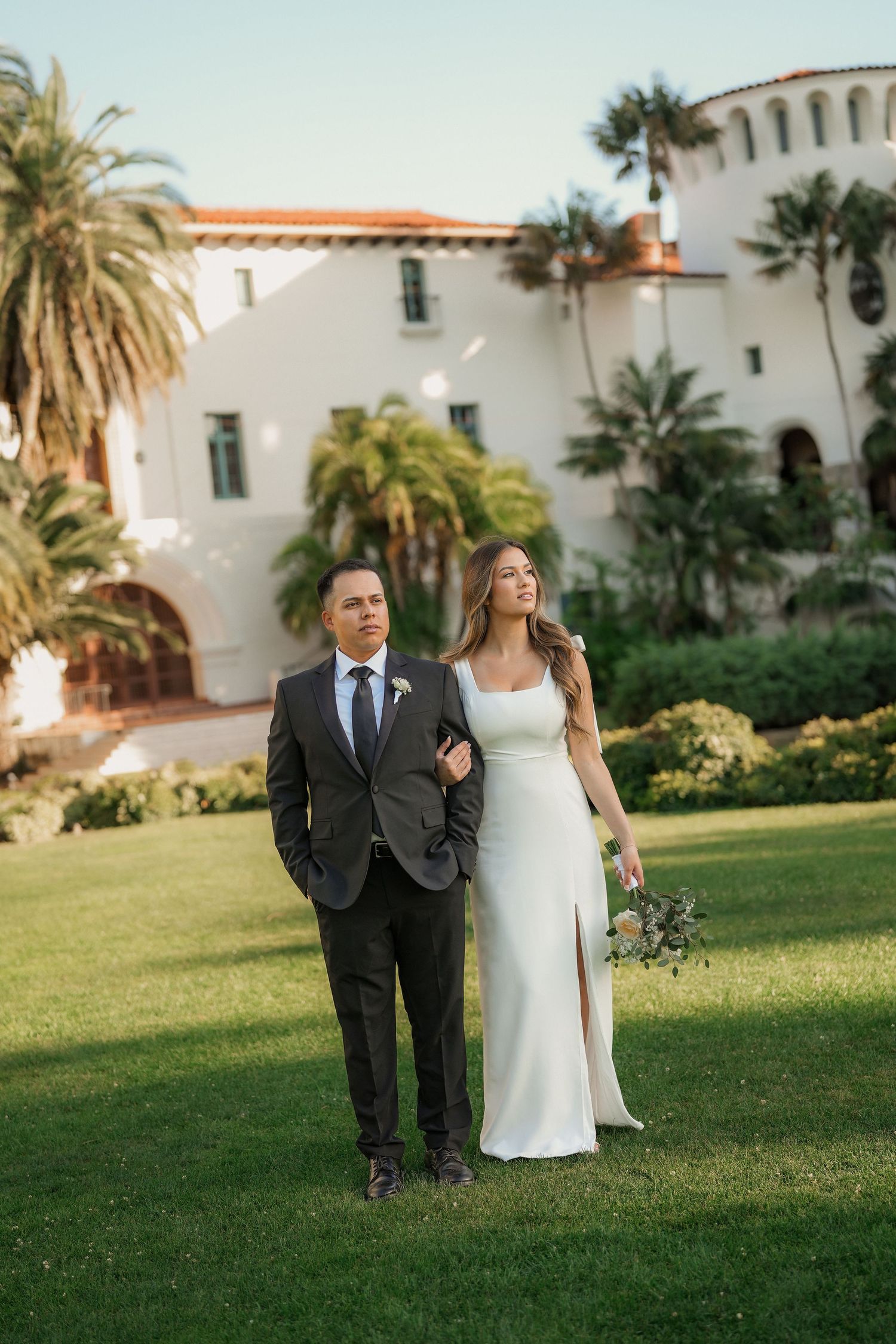 Bride in a white gown and groom in a gray suit pose arm in arm on a green lawn in front of a Spanish-style building.