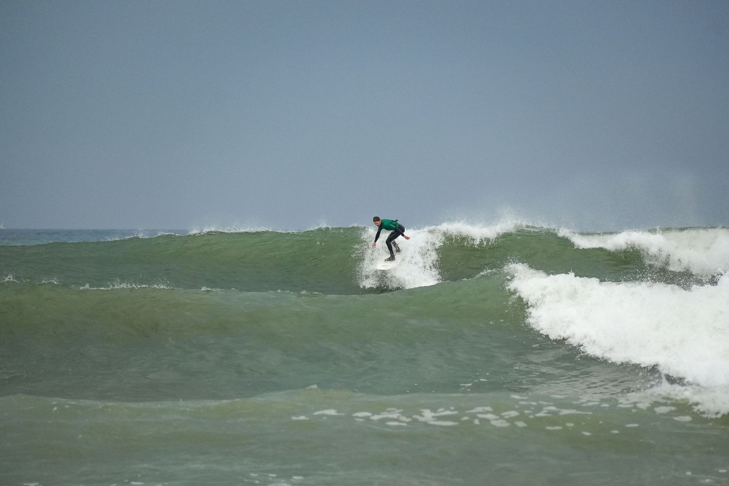 Surfer in a teal wetsuit carves along the face of a large green ocean wave as whitewater crashes nearby.