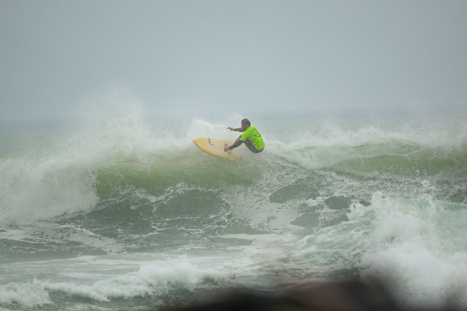 Surfer in a neon green shirt performs a sharp turn on a yellow surfboard in rough, churning waves.
