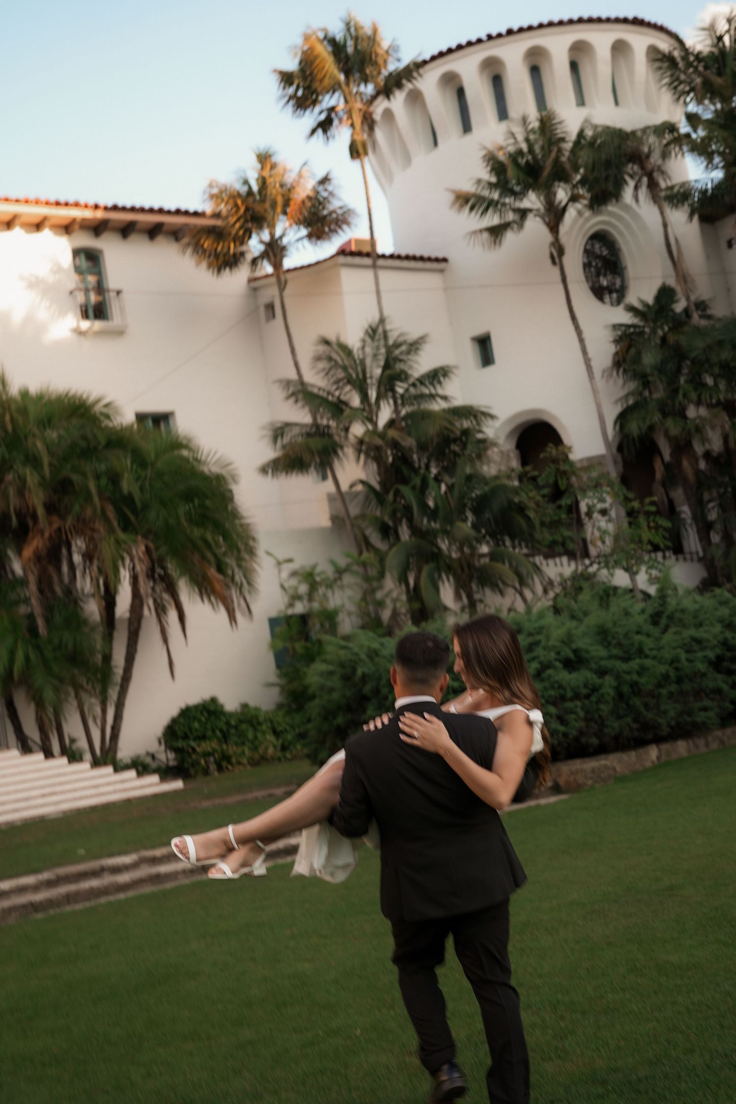 Groom carries his bride across a green lawn toward a whitewashed Spanish-style building surrounded by palm trees and lush plants.