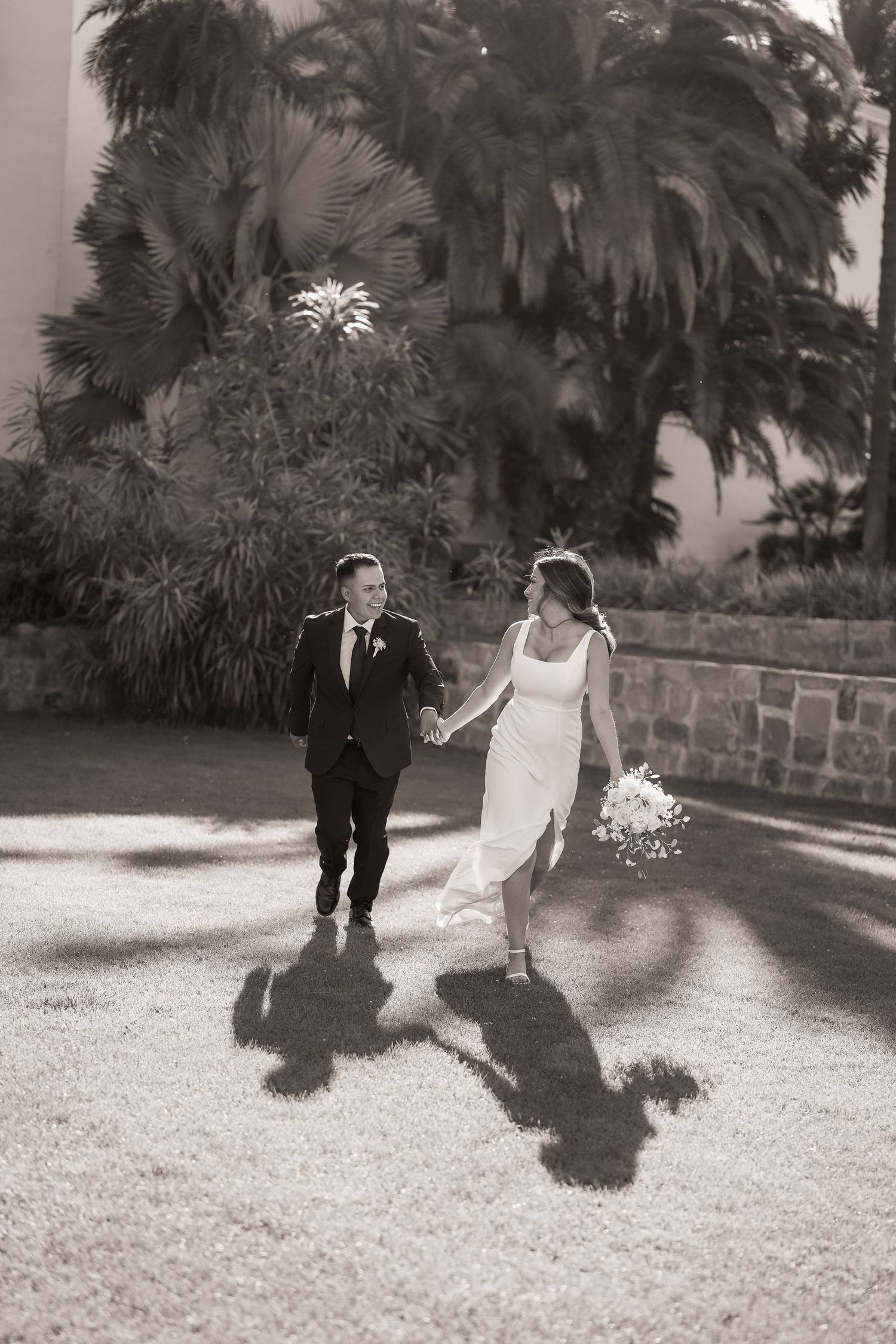 Black and white photo of a bride and groom holding hands and walking across a lawn beside palm trees.