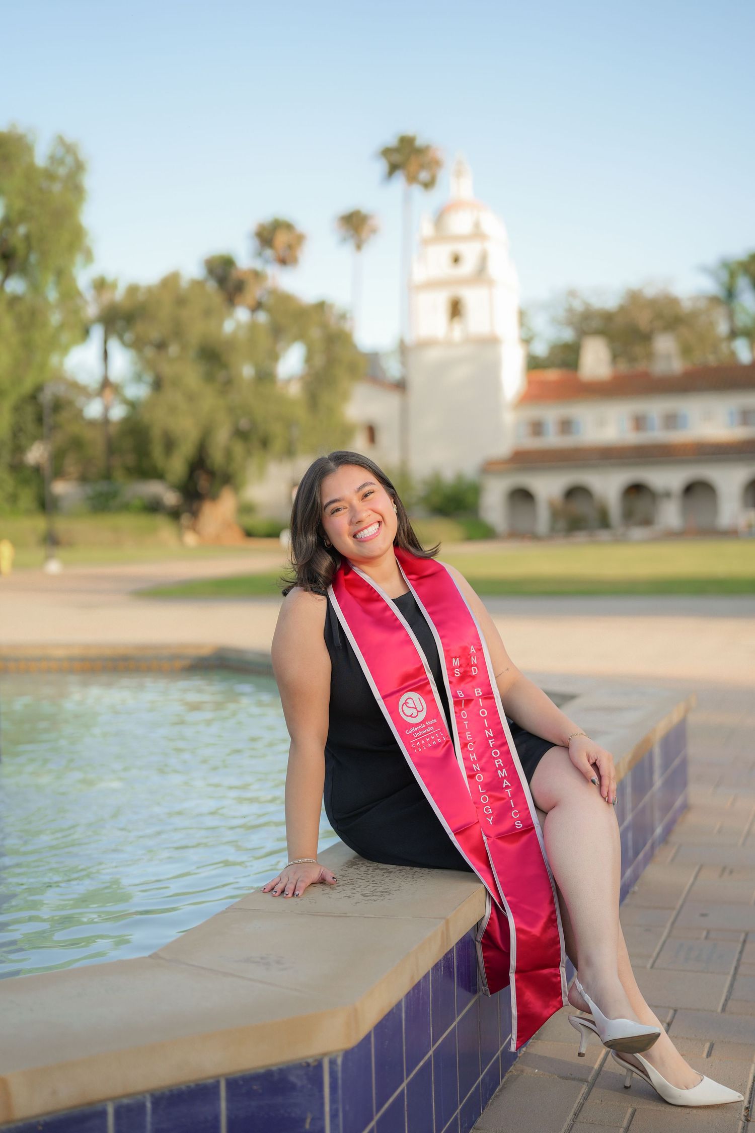 Smiling graduate in a black dress and red sash sits on a tiled fountain ledge with a white bell tower behind her.