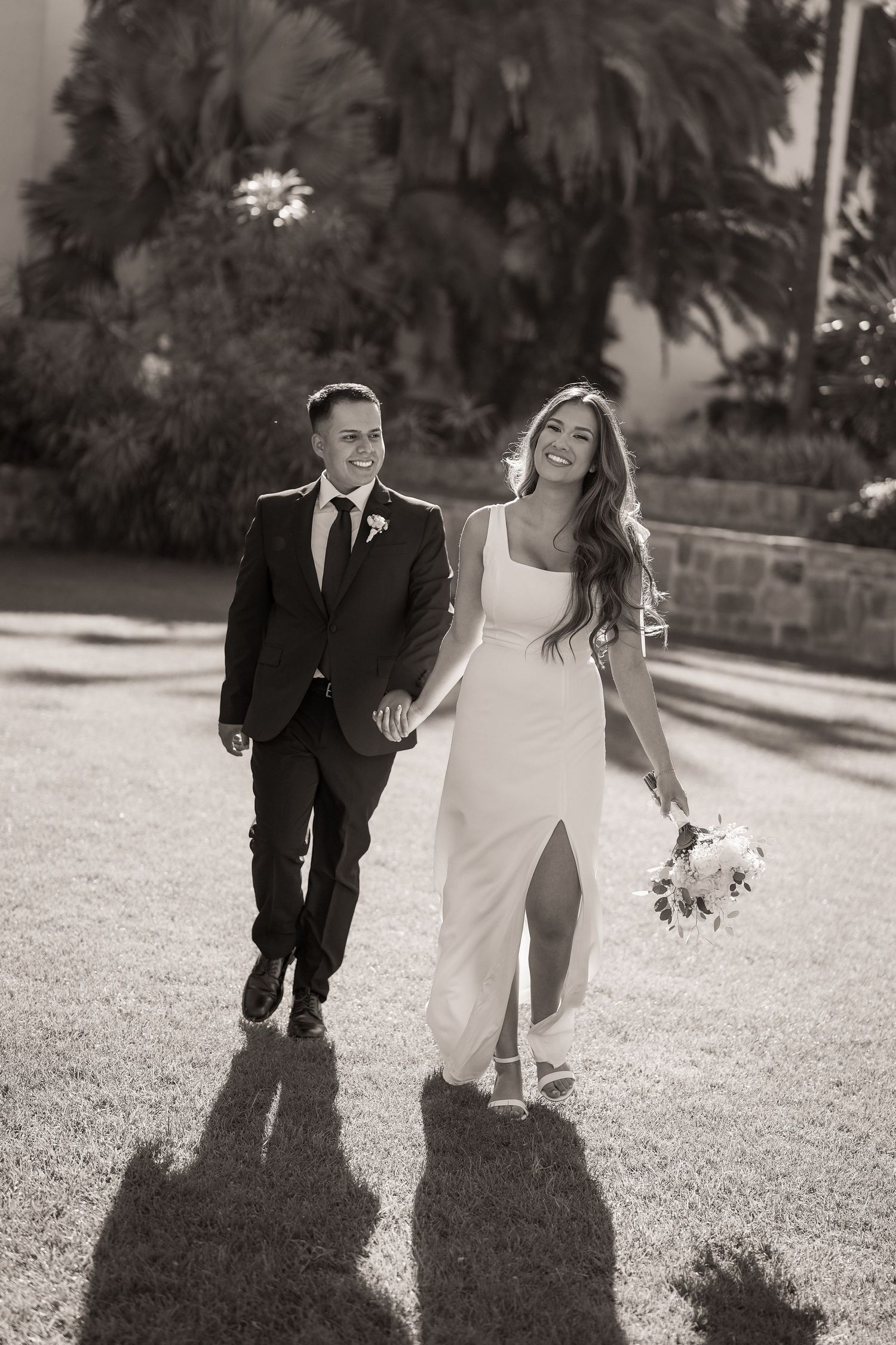 Black and white photo of a smiling couple holding hands, the bride in a slit gown with a bouquet, walking on a lawn.
