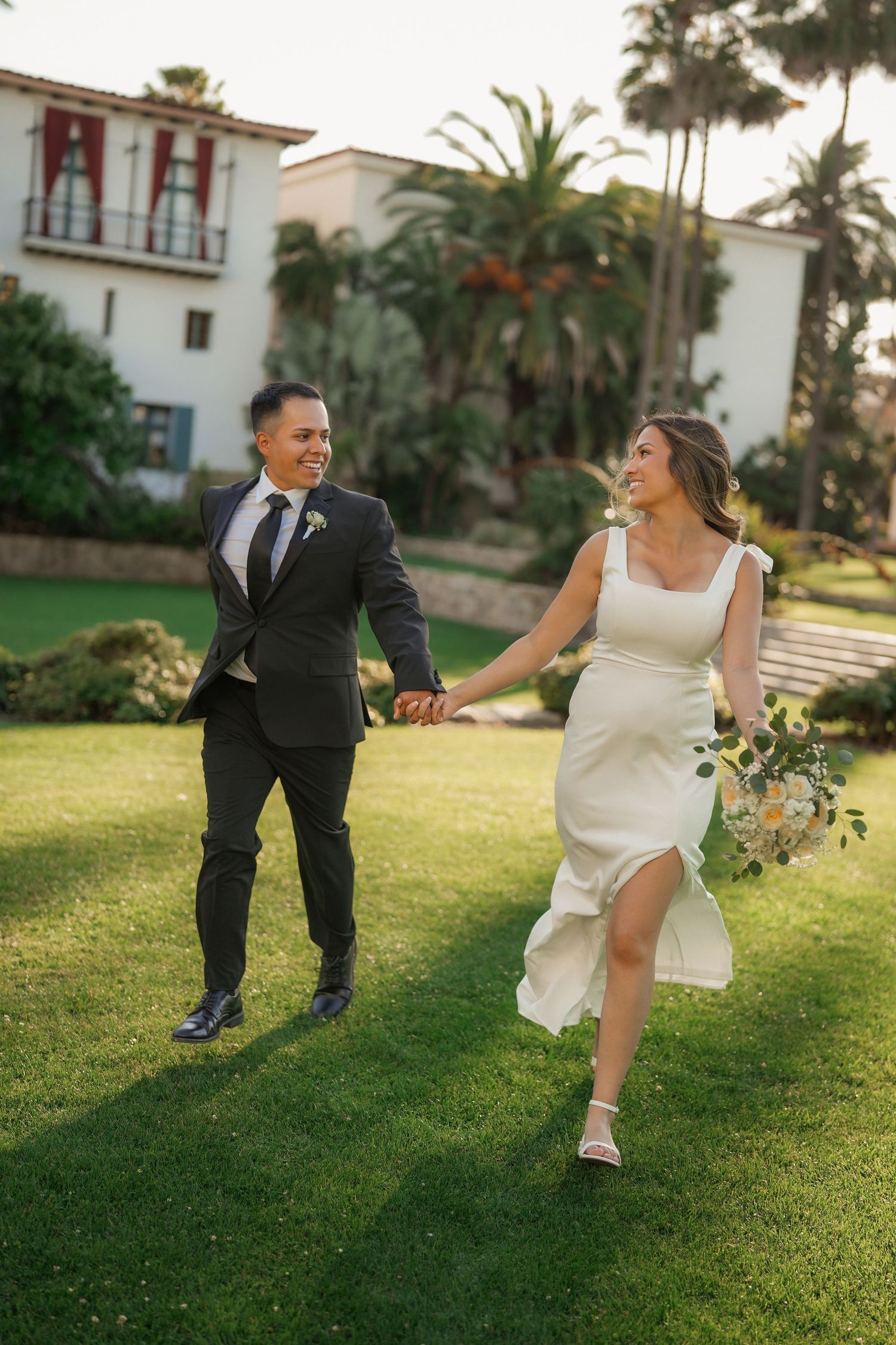 Laughing bride holding a bouquet runs hand in hand with her groom across a grassy lawn at golden hour.