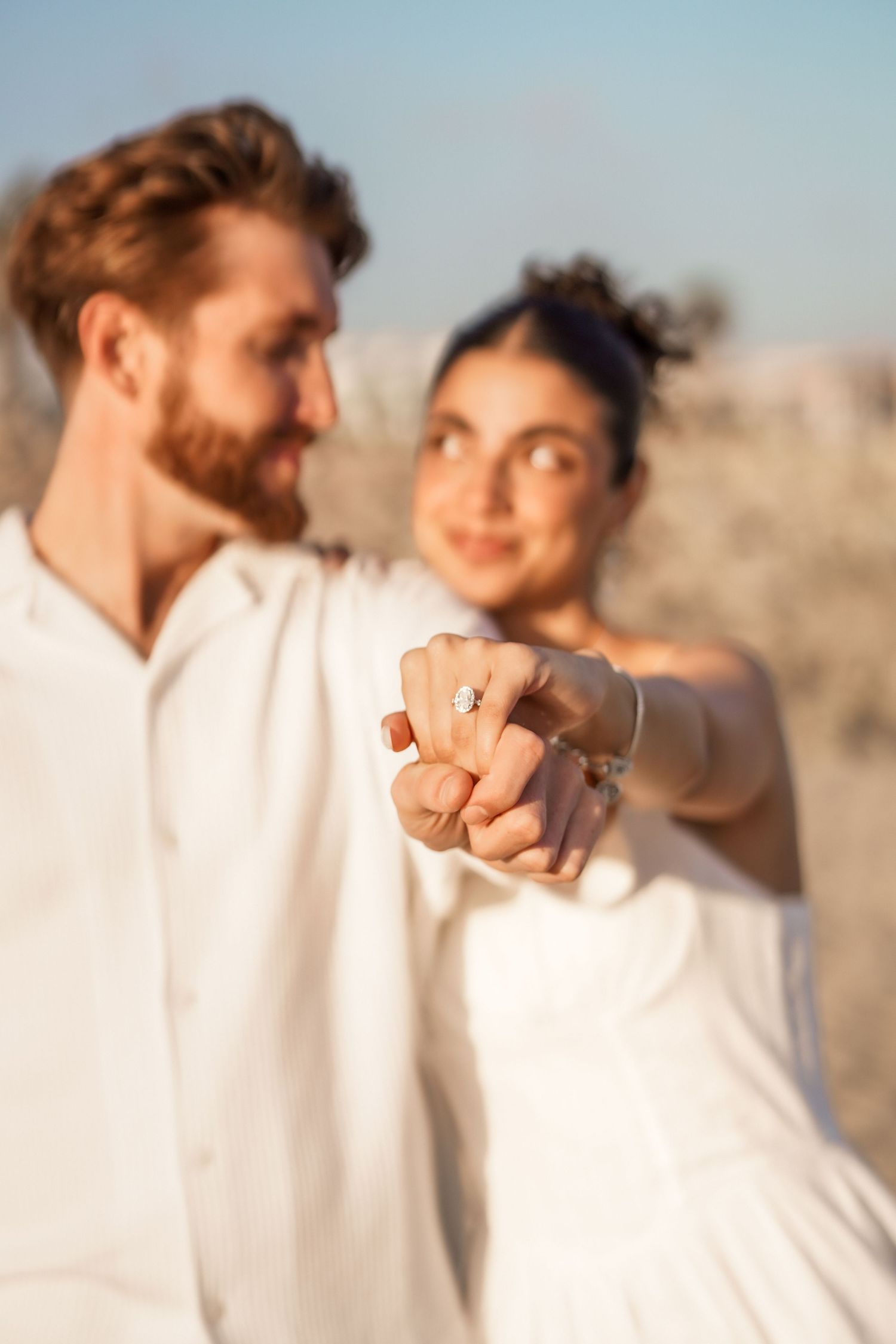 Close-up of a couple's hands showing a sparkling engagement ring, with the smiling pair softly blurred behind.