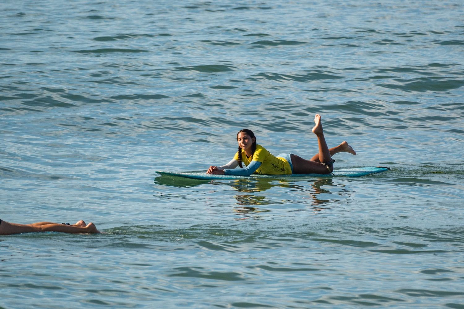 A surfer in a yellow shirt lies prone on a blue longboard paddling on calm ocean water.