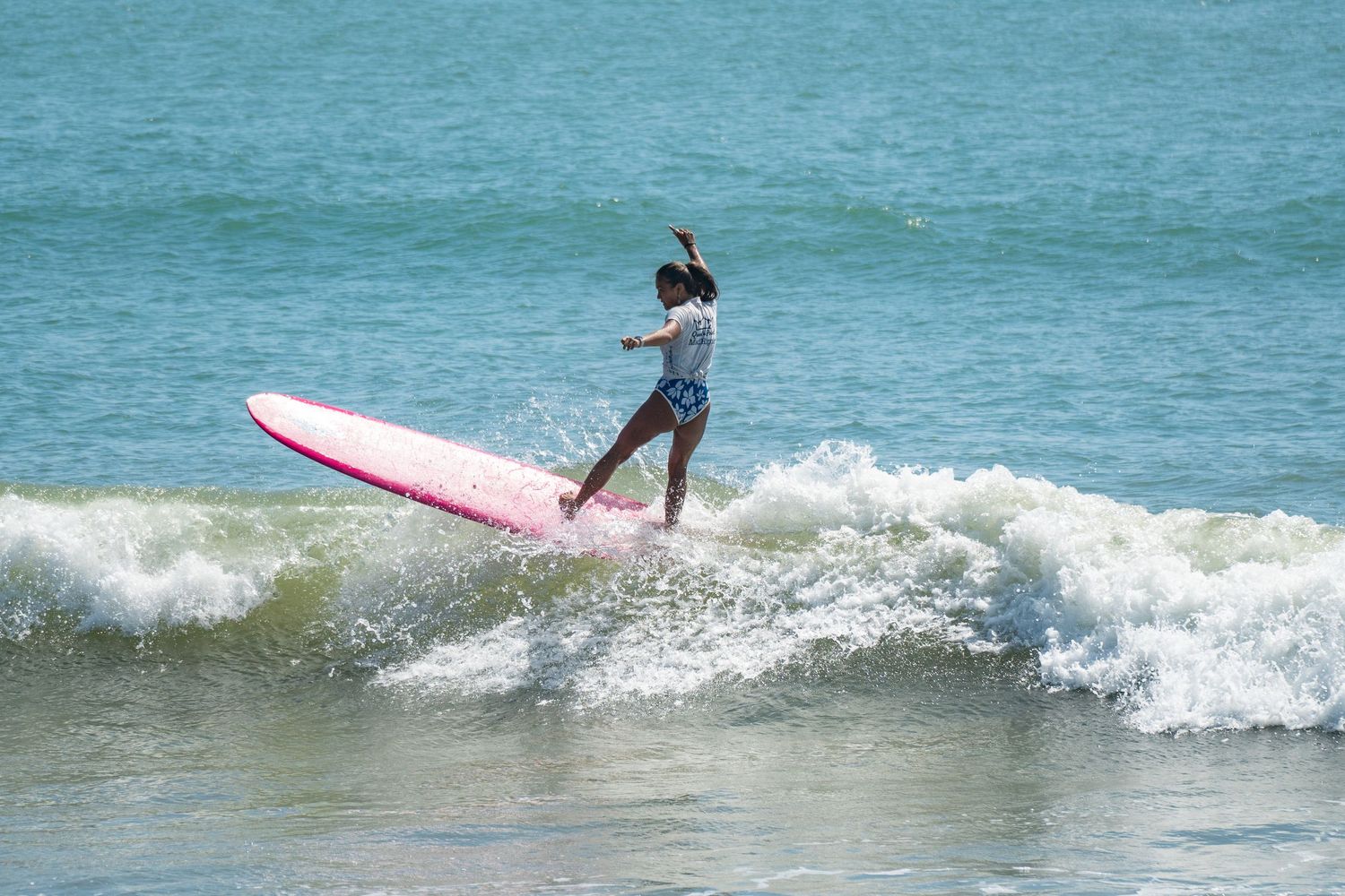 A surfer performs a turn on a pink longboard atop a crashing turquoise wave with one arm raised.