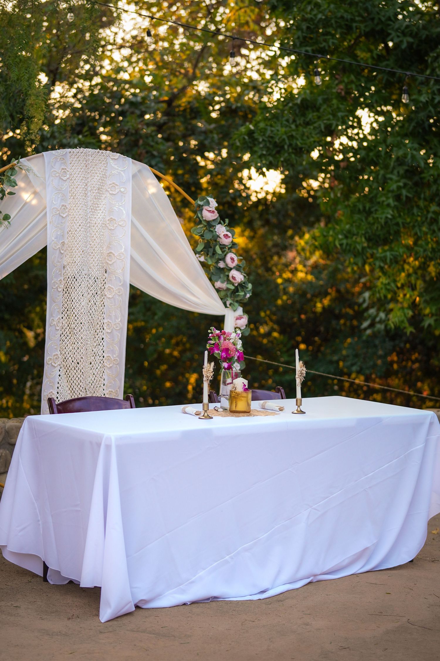 Outdoor wedding ceremony table draped in white linen with candles, pink floral arrangements, and a macrame arch backdrop among trees.