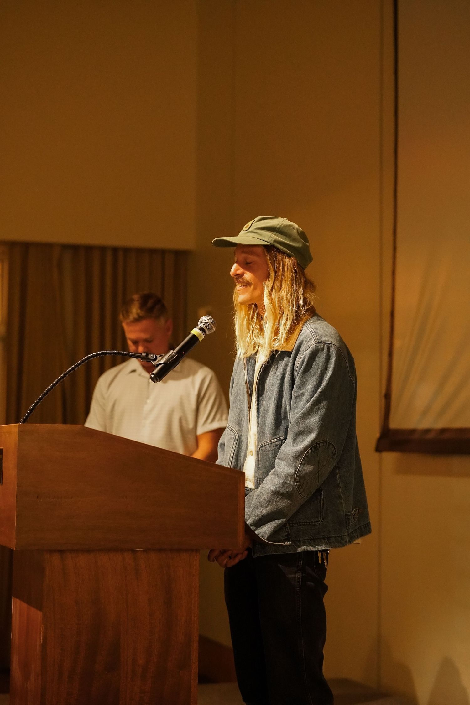 Person in a green cap and denim jacket smiles at a wooden podium microphone while another person sits behind it during a program.