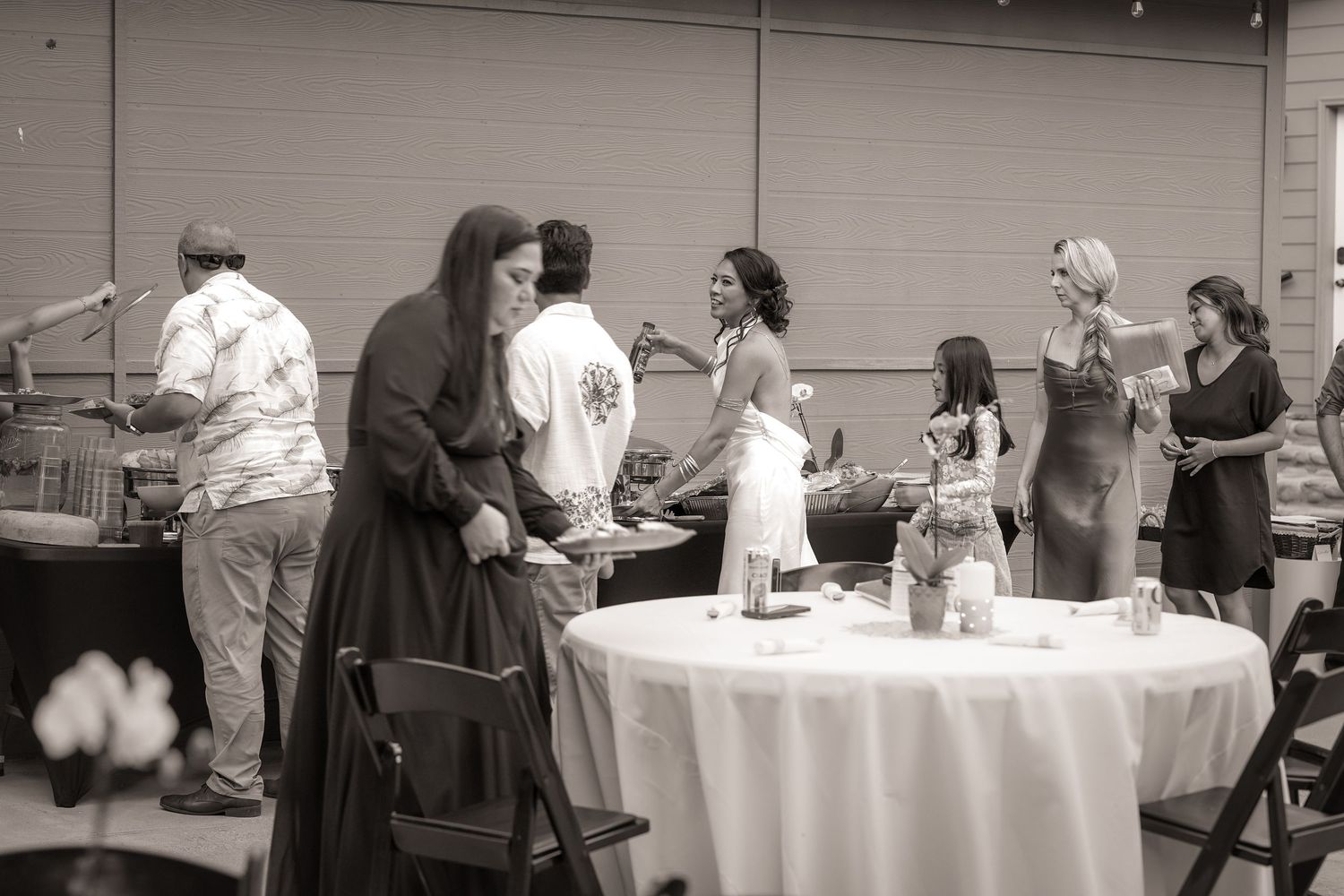 Black and white photo of wedding guests mingling and serving themselves near a buffet table at an outdoor reception.