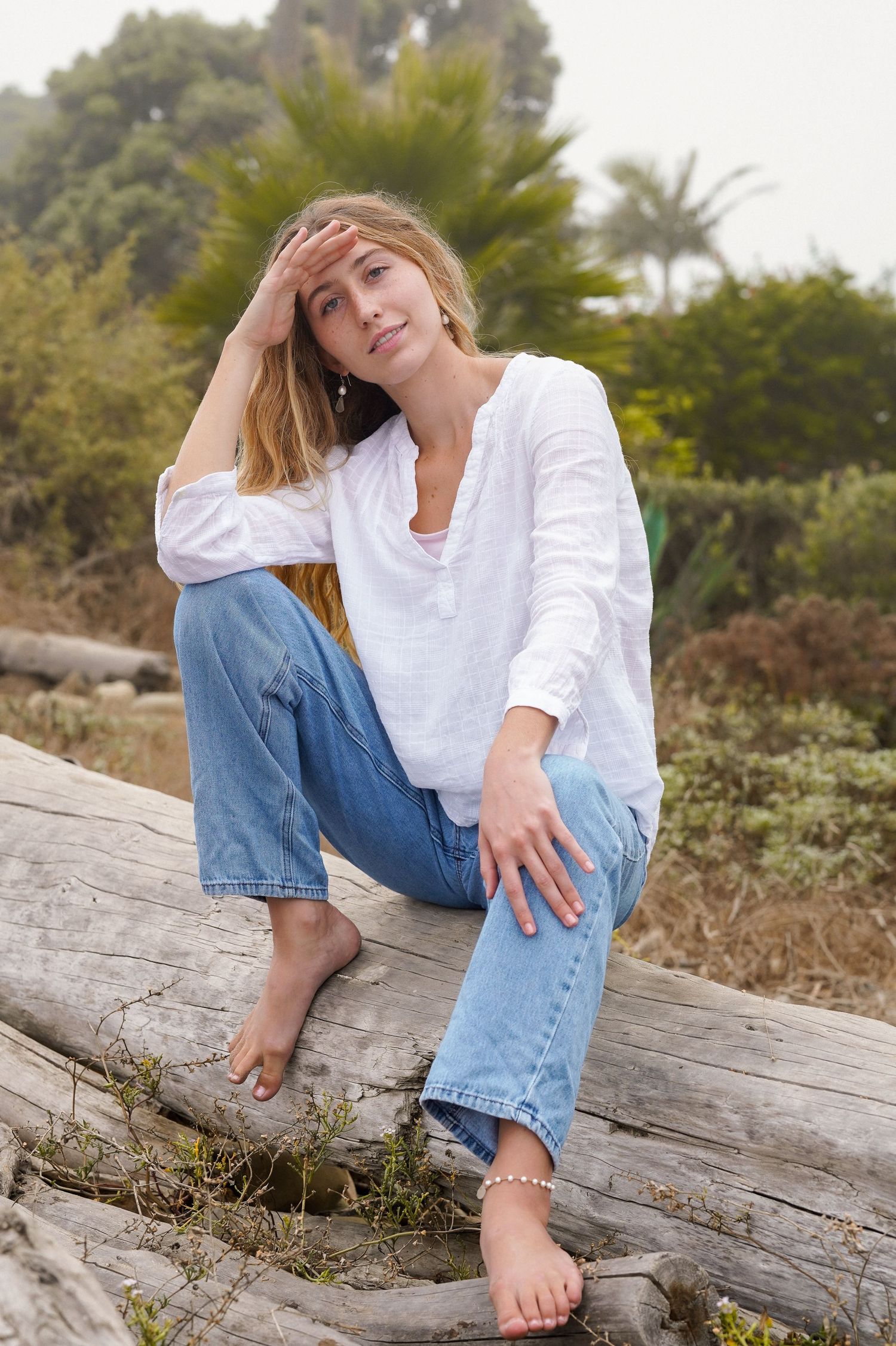 Barefoot woman in a white linen shirt and jeans sits on a driftwood log with palm trees in the background.
