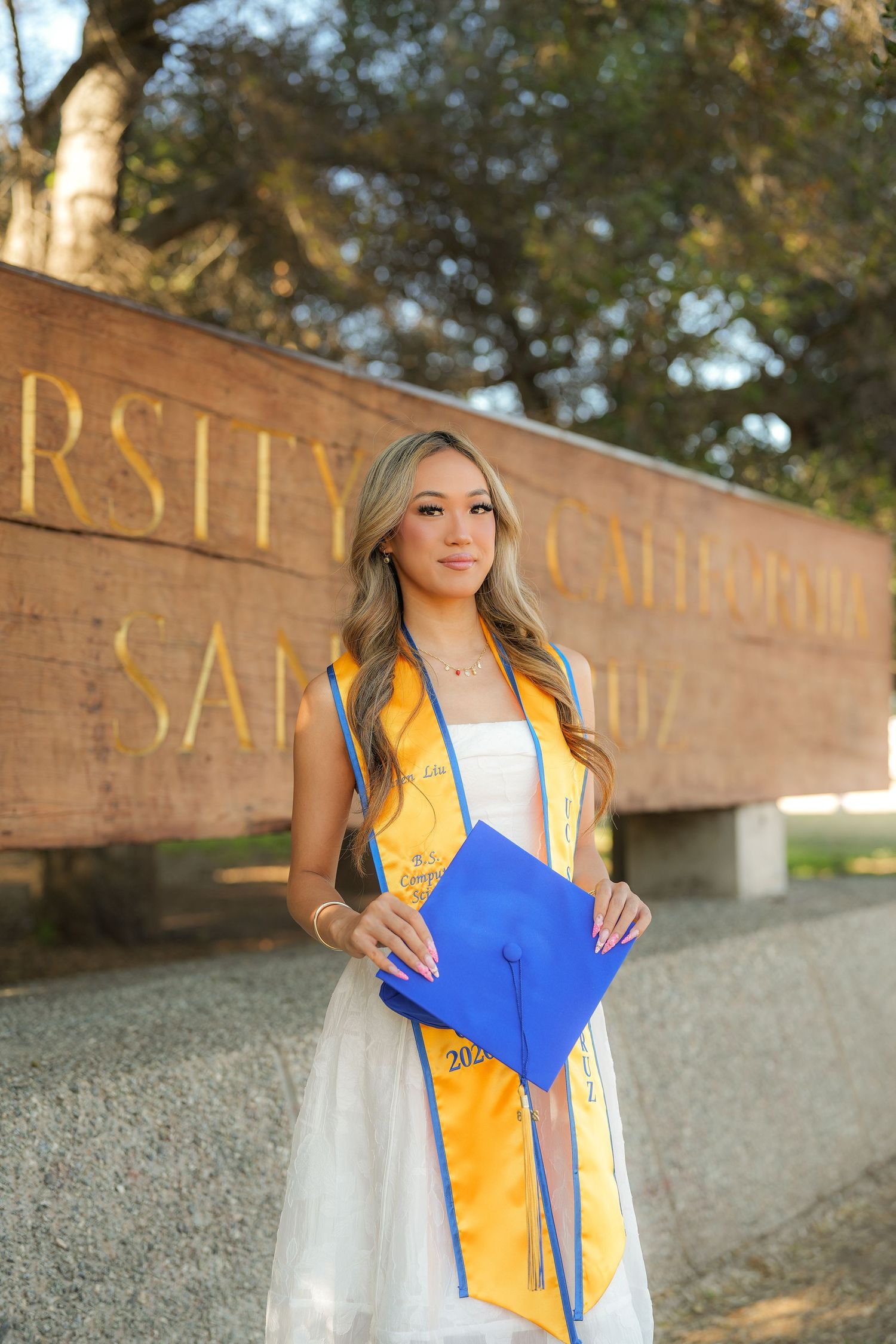 Graduate in a white dress and gold sash holds a blue cap beside a wooden university sign with gold lettering and trees behind.