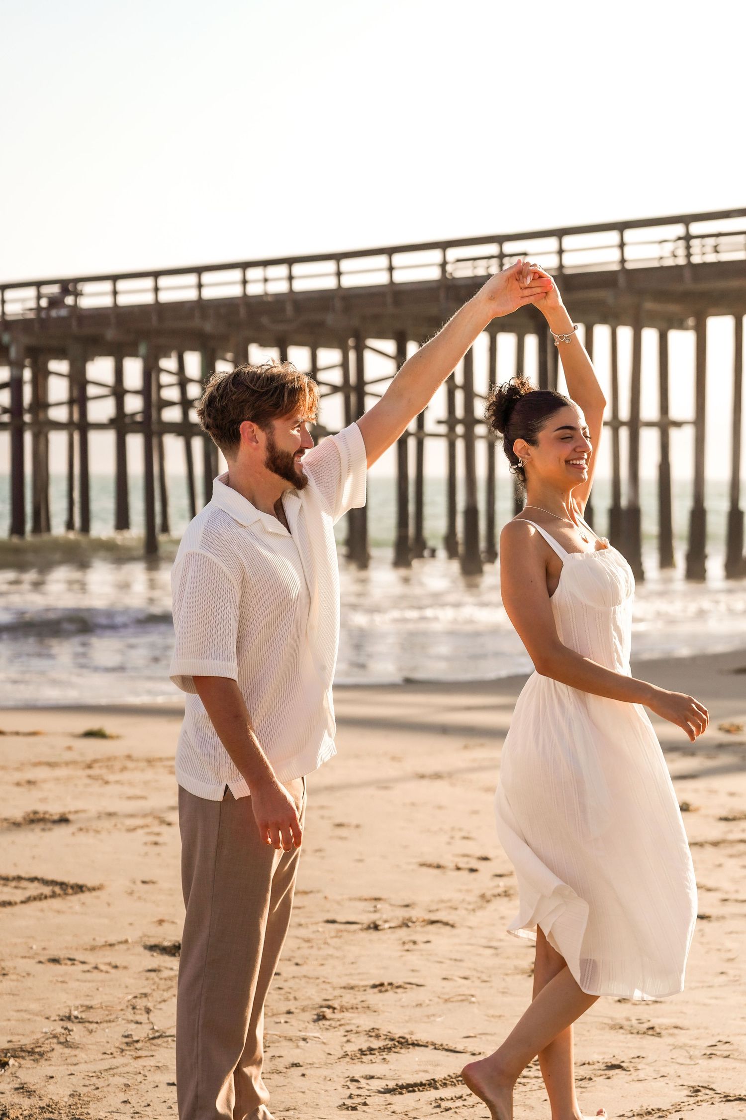 Smiling couple dances on the beach as the man twirls his partner in a white dress, with a wooden pier behind them.