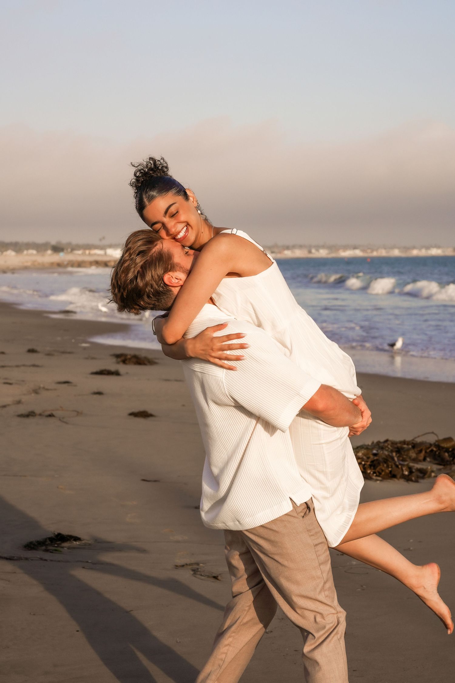 Couple in white and khaki laughing as he lifts her in an embrace on a sandy beach at golden hour.