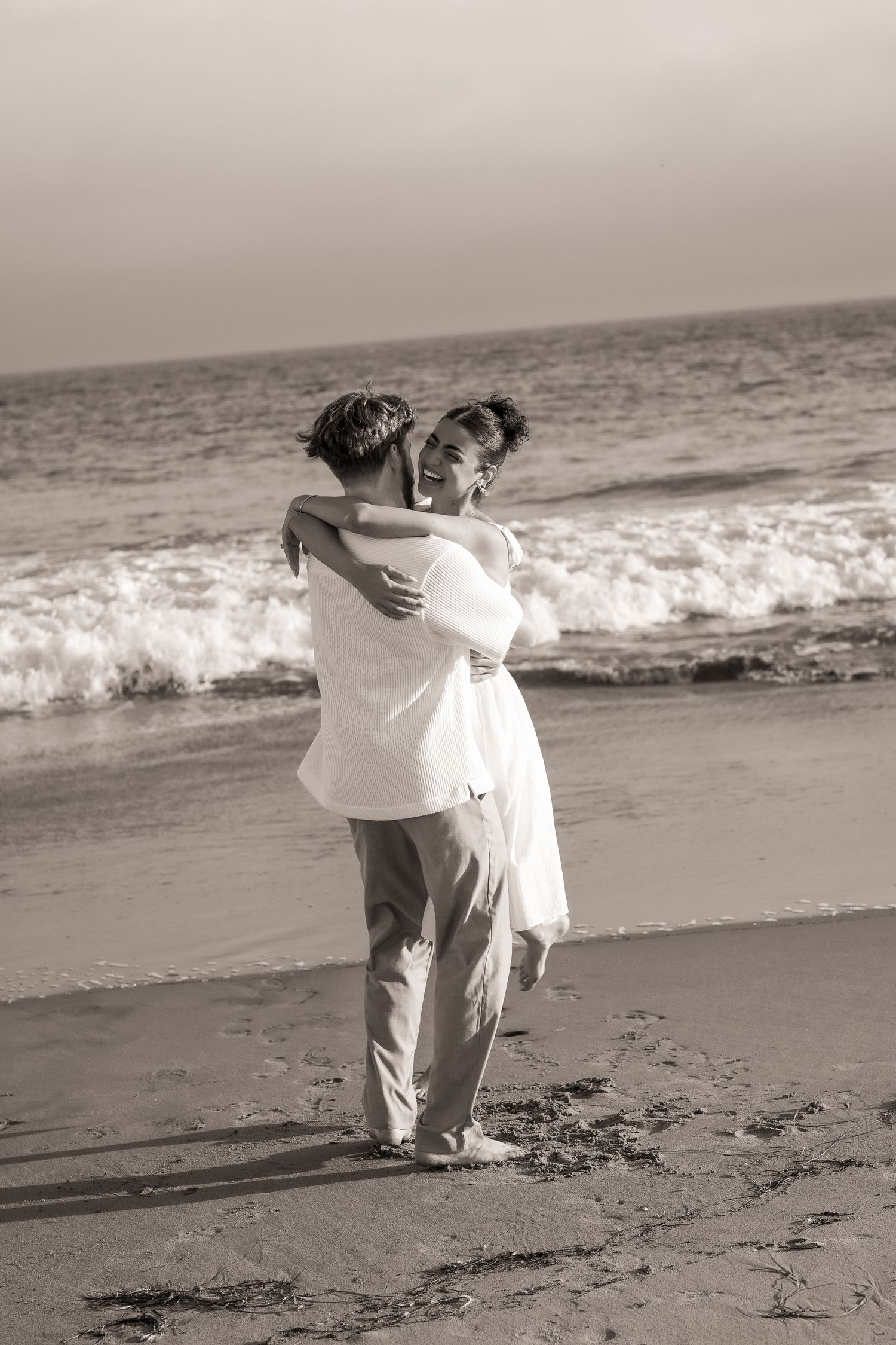 Black and white photo of a couple embracing and laughing on the beach as ocean waves roll in behind them.