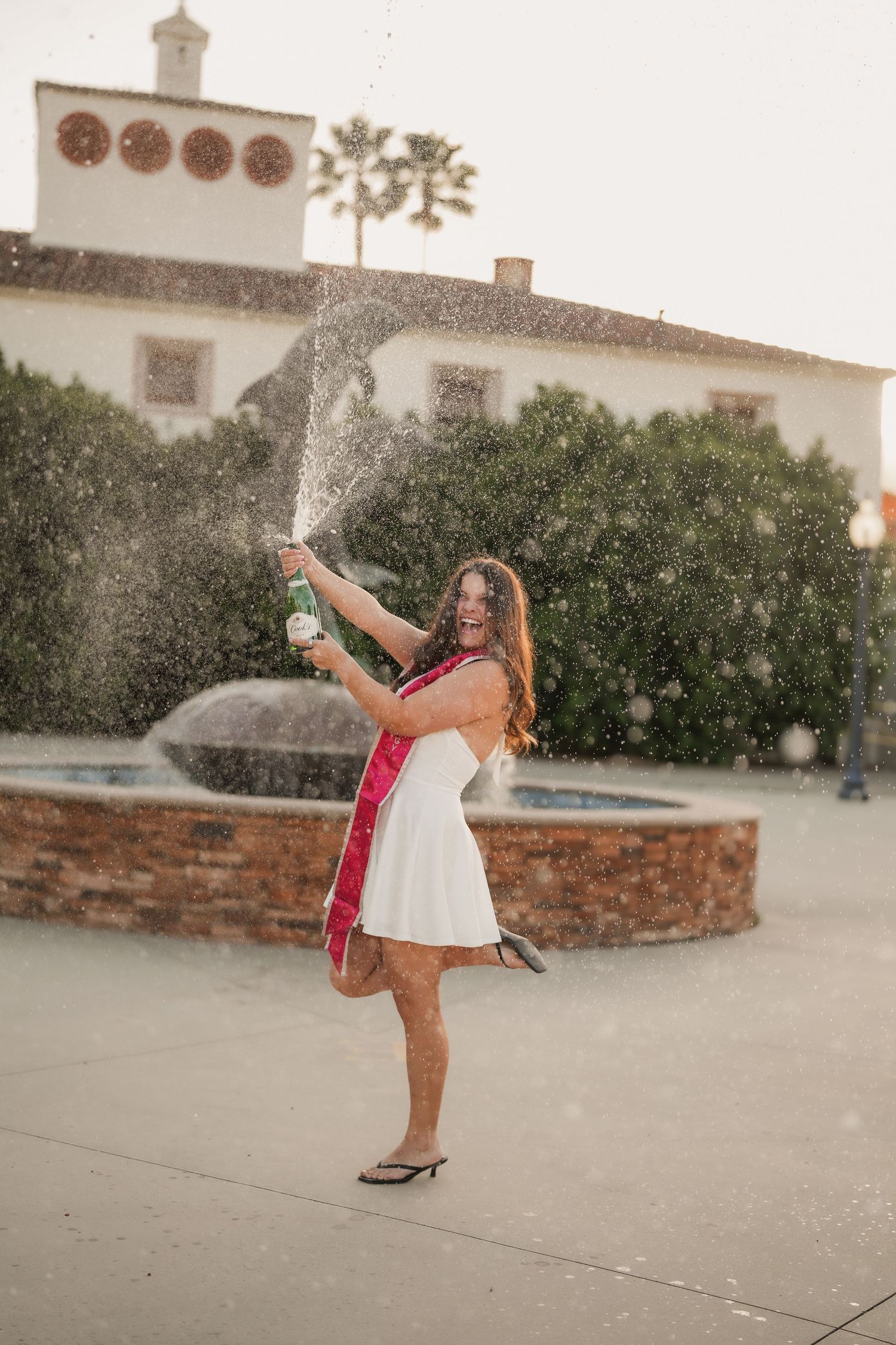 A graduate in a white dress and red sash laughs as she sprays champagne in front of a brick fountain on a sunny campus.