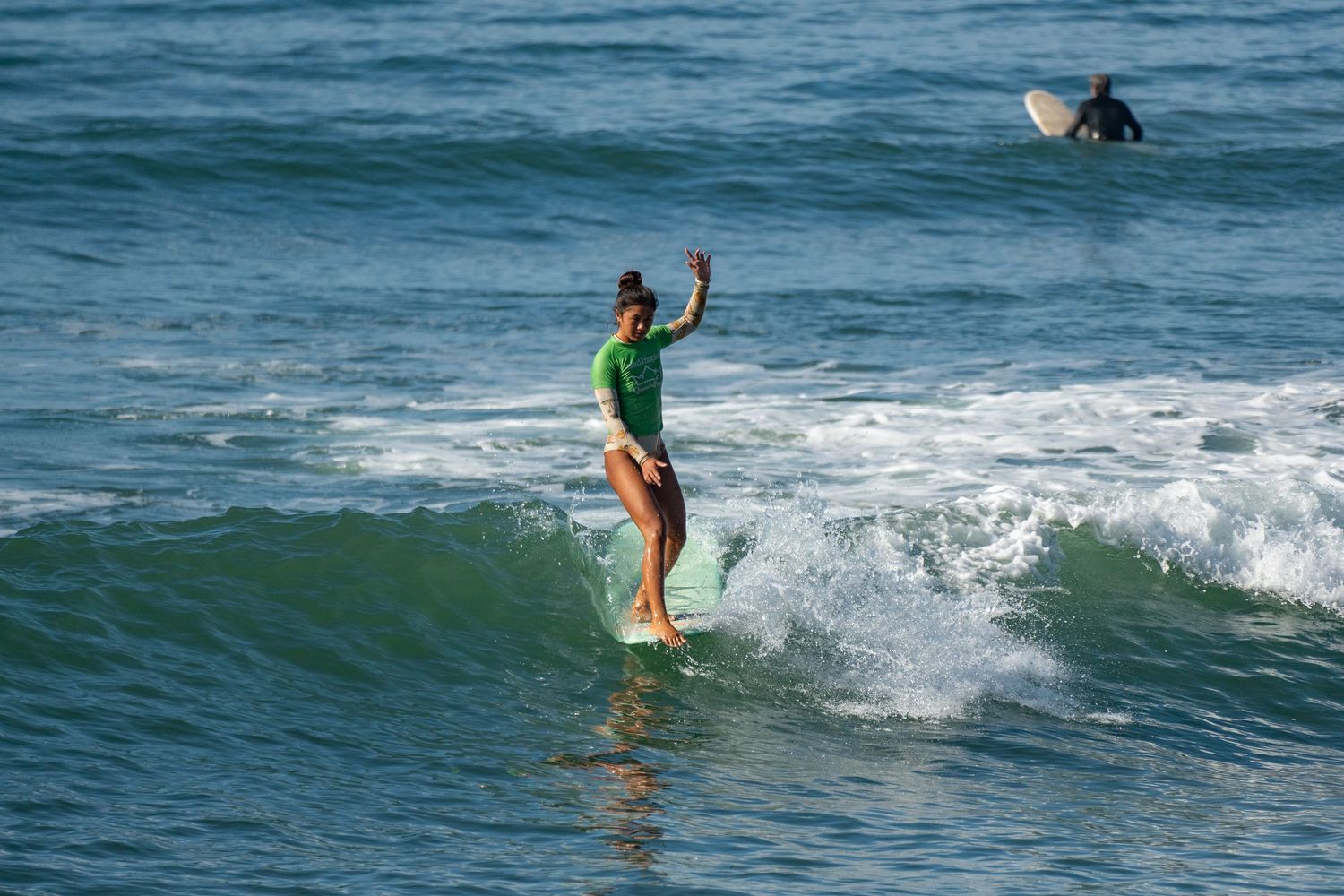 A longboarder in a green top raises one arm while riding a green ocean wave as another surfer waits behind.