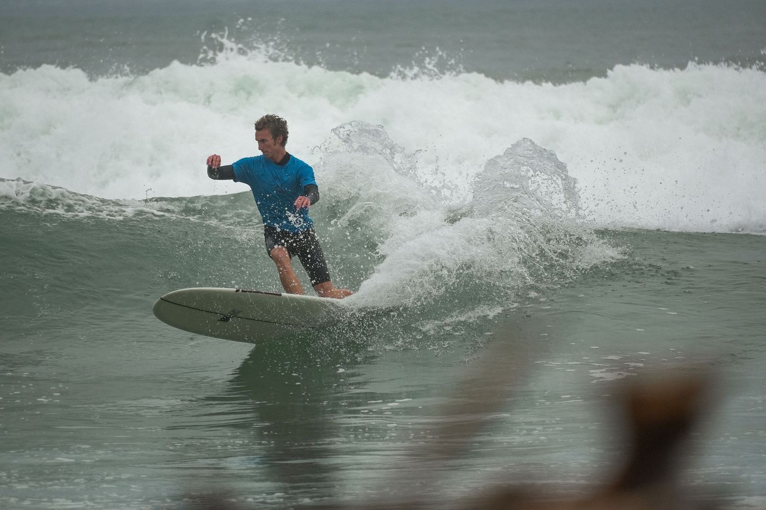 Surfer in a blue shirt and patterned shorts leans into a turn on a white longboard in choppy surf.