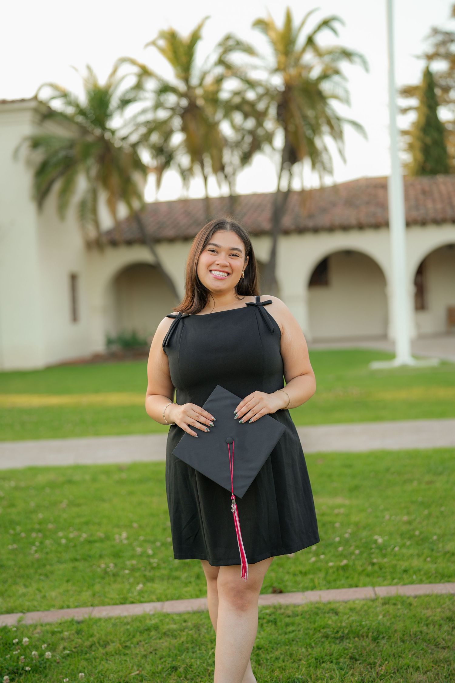 A smiling graduate in a black dress holds her cap with a red tassel on a grassy lawn in front of palm trees and arched buildings.