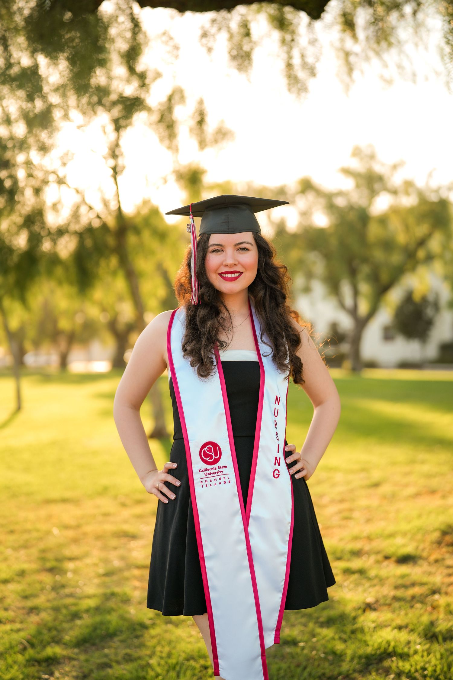 Smiling nursing graduate wearing a cap, black dress and white sash poses with hands on hips on a sunlit grassy lawn.