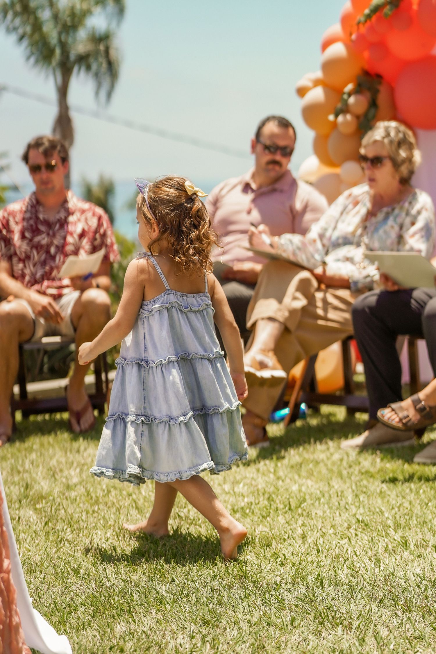 Barefoot toddler in a blue tiered dress walks across a sunny lawn at an outdoor party as guests sit nearby beside an orange balloon garland.