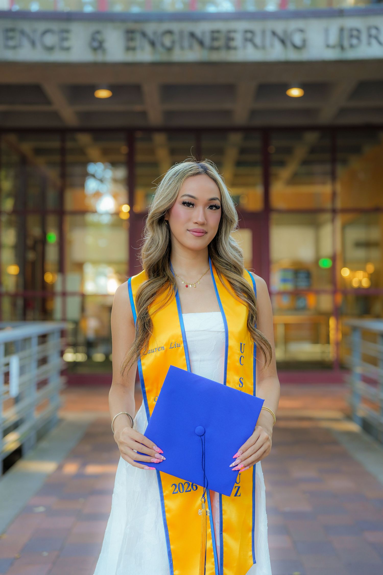Smiling graduate in a white dress, gold stole and 2026 sash holds a blue cap in front of the Science and Engineering Library.