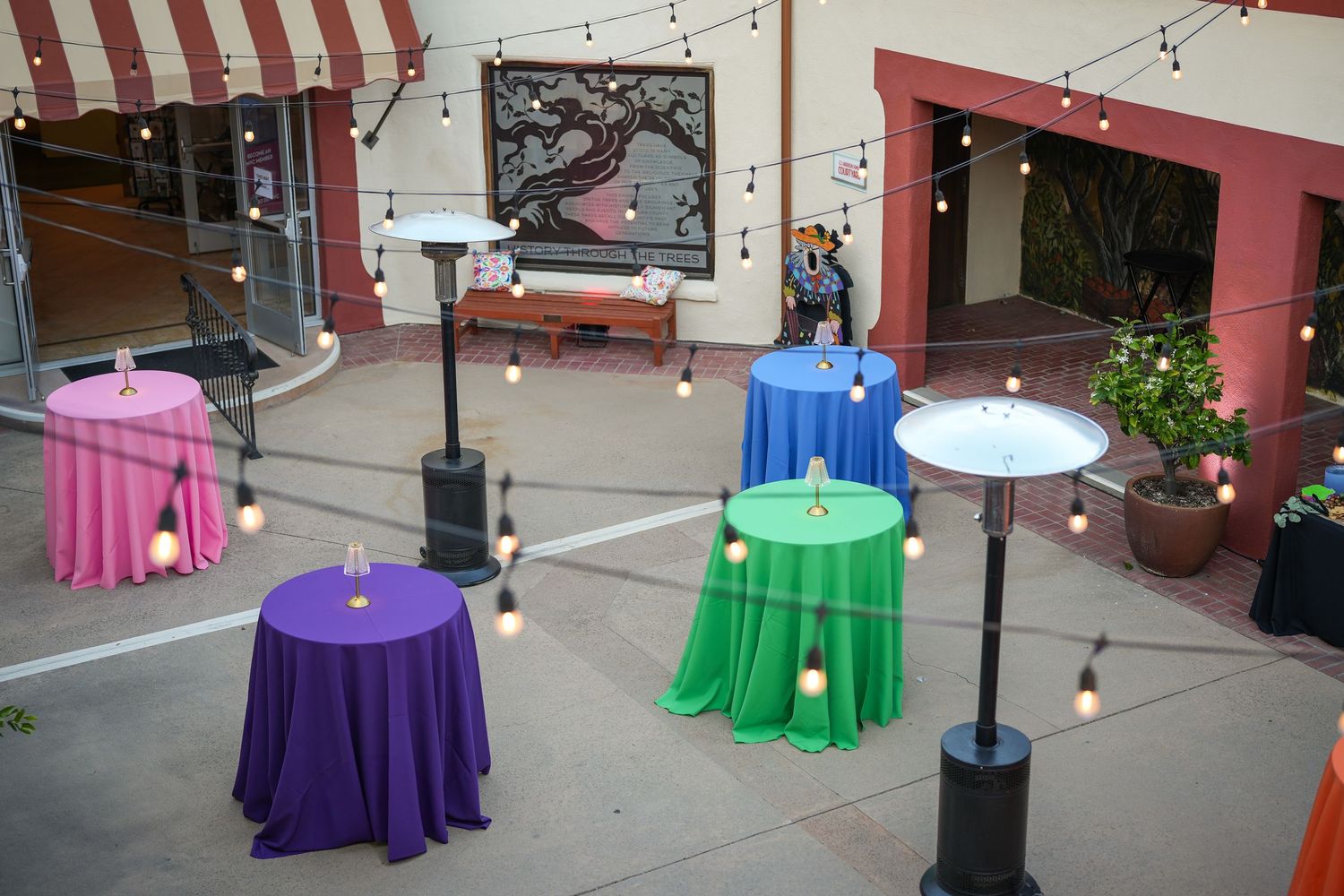 Overhead view of purple, green, pink and blue draped cocktail tables with string lights and heaters in a festive courtyard.