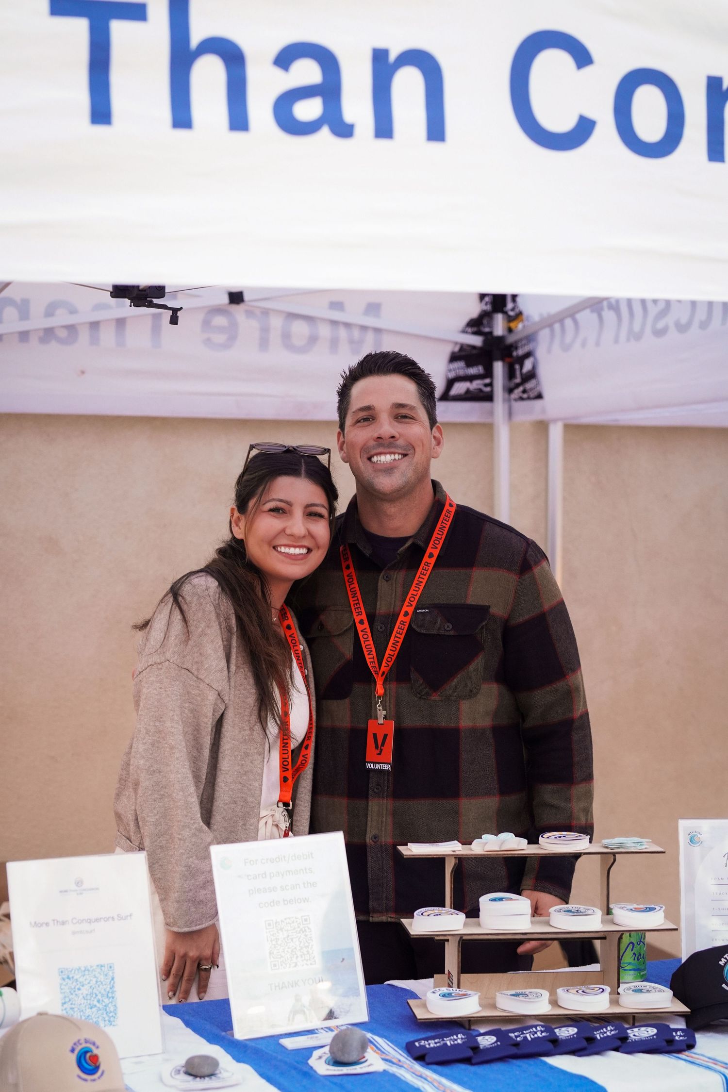 A smiling man and woman wearing lanyards stand together at a More Than Conquerors booth table with QR code signs and displays.