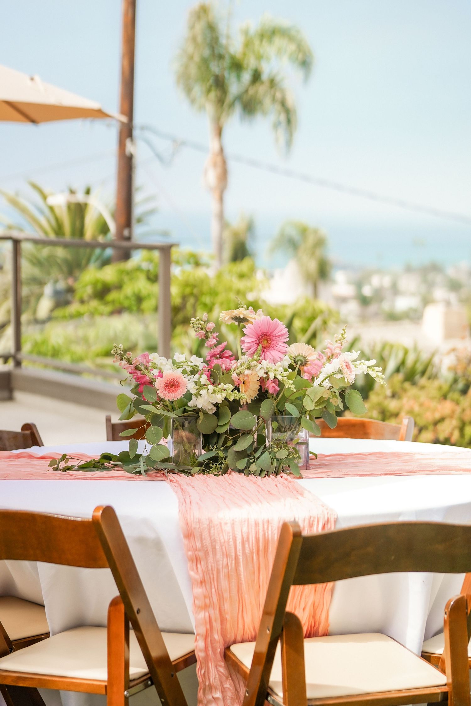 Pink gerbera daisy and greenery centerpiece on a white table with coral runner and wooden chairs, set against palm trees and an ocean view.