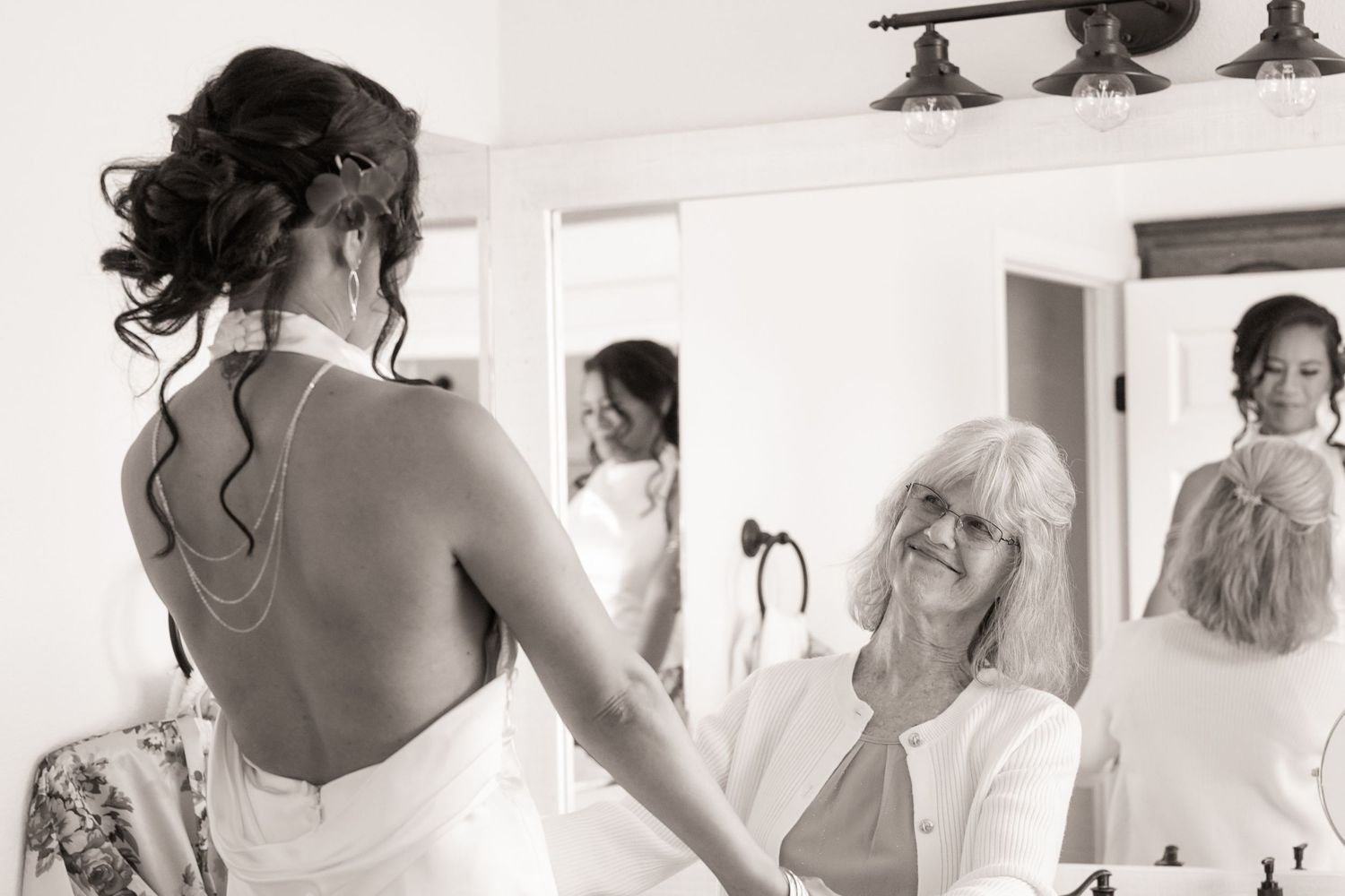 Bride in a backless gown with an elegant updo looks in the mirror as a smiling older woman watches during wedding preparations.