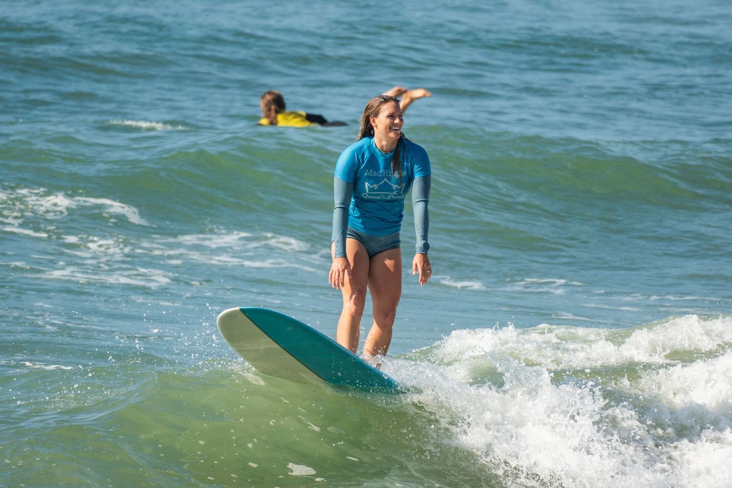 A smiling surfer in a blue wetsuit top stands on a teal longboard in whitewater as a surfer paddles behind.